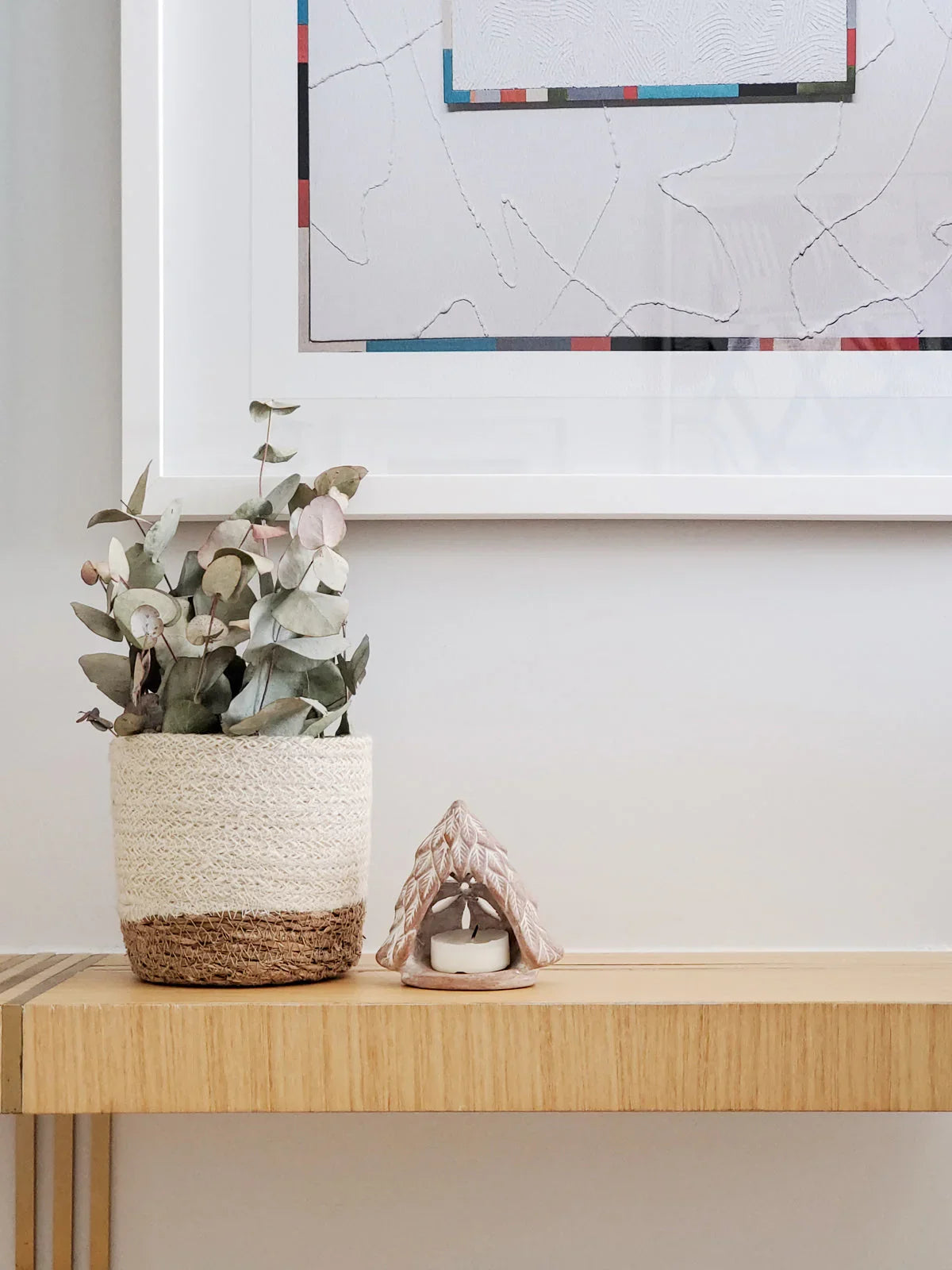 Terracotta candle holder shaped like a tree with a tealight candle on wooden console table beside woven planter with eucalyptus