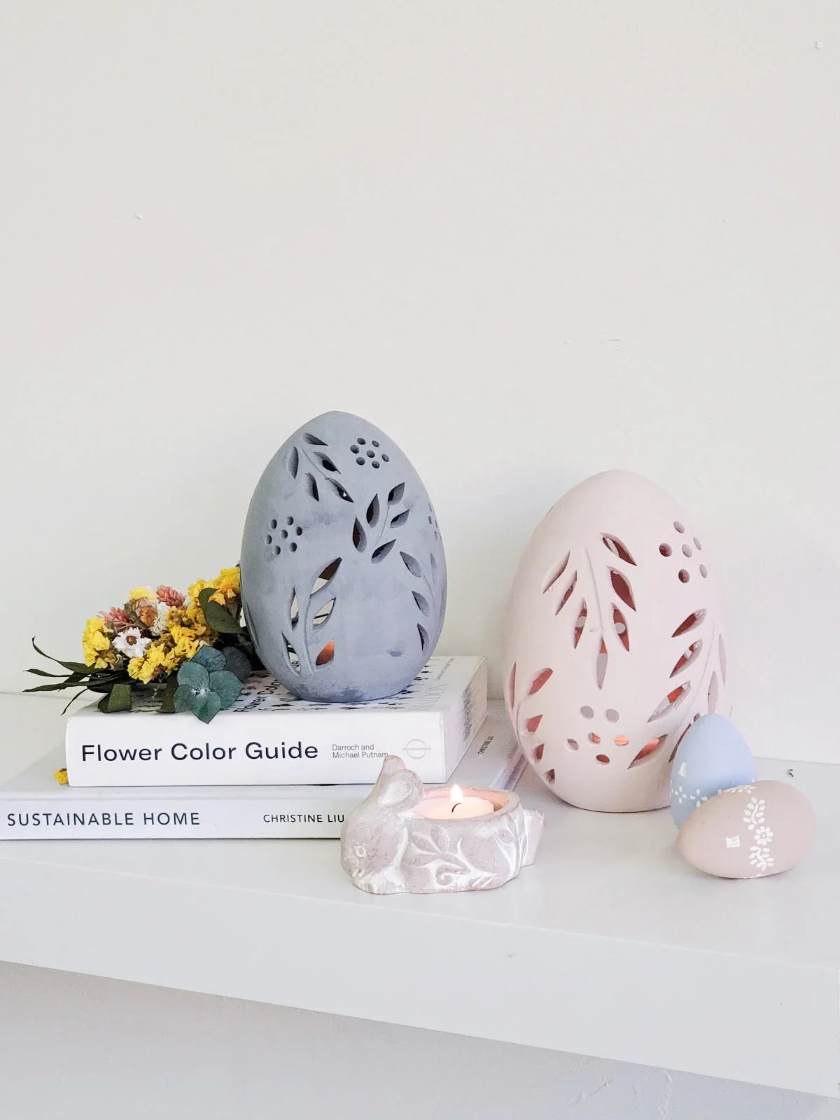 Decorative terracotta egg-shaped candle lanterns with floral cutouts on a white shelf with books and small candles