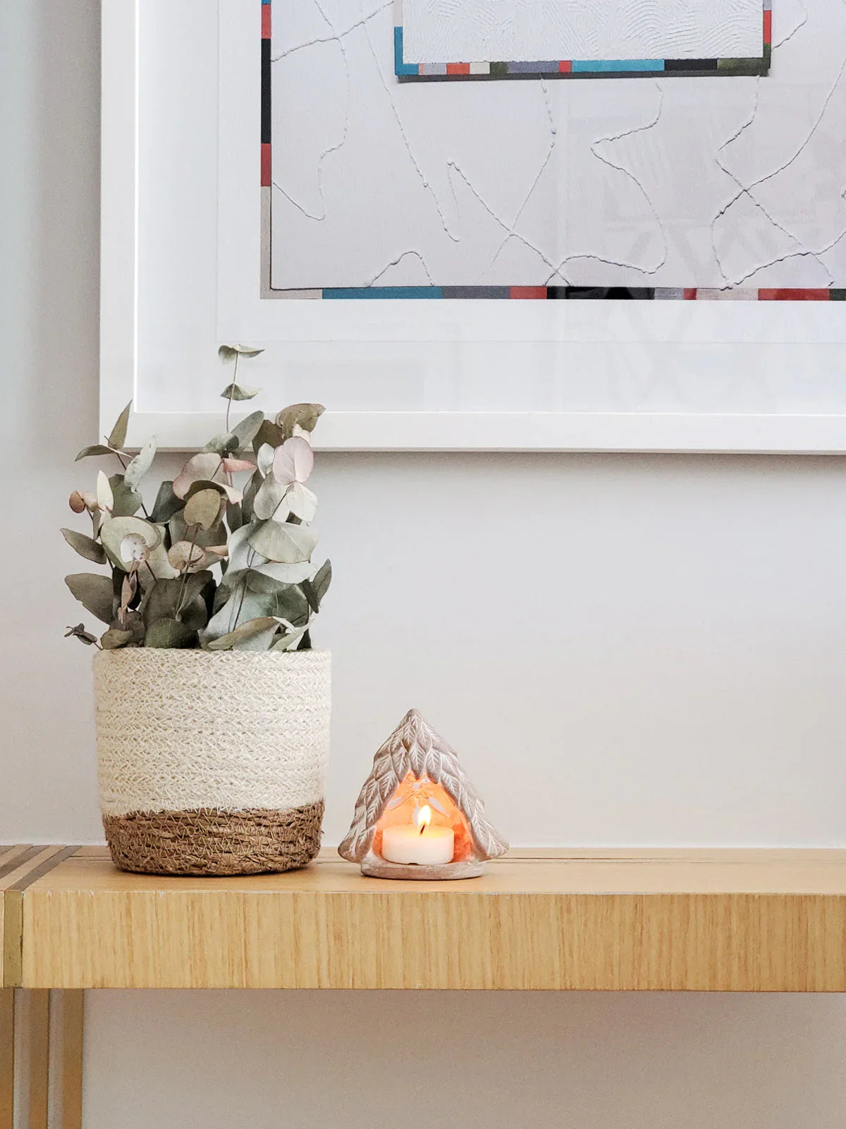Terracotta tree-shaped candle holder with lit tea light on wooden table next to woven plant pot with eucalyptus