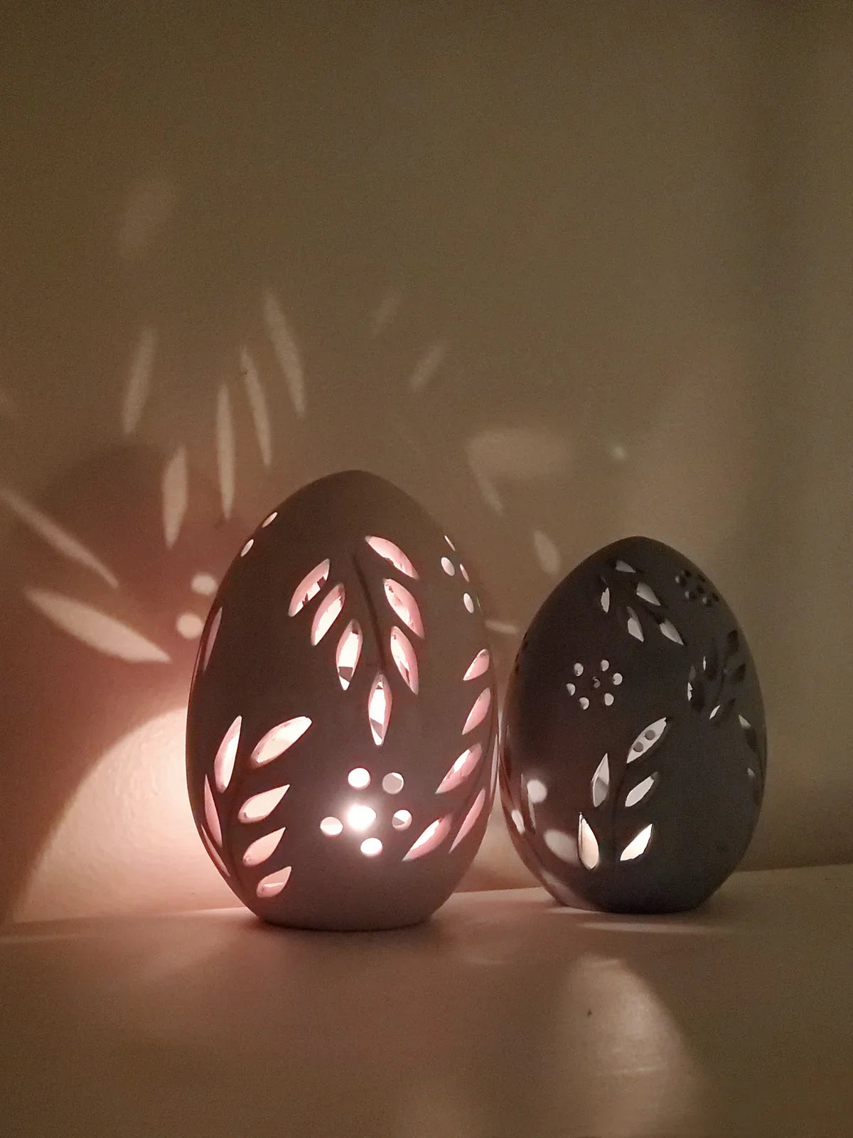 Two egg-shaped terracotta candle lanterns with leaf and flower cutouts casting soft shadows on the wall