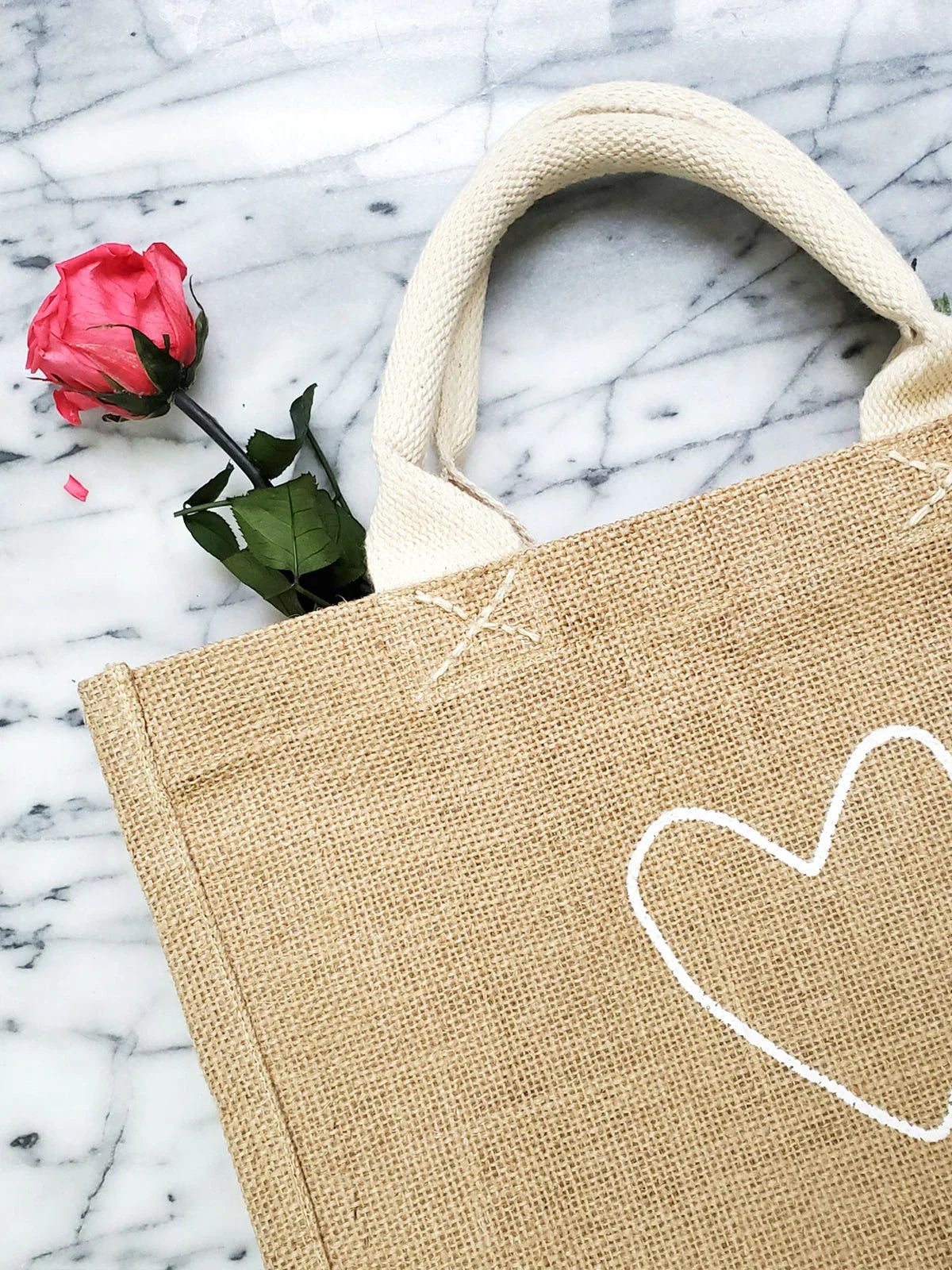 Natural jute tote bag with white heart print and sturdy cream handles on marble surface with pink rose