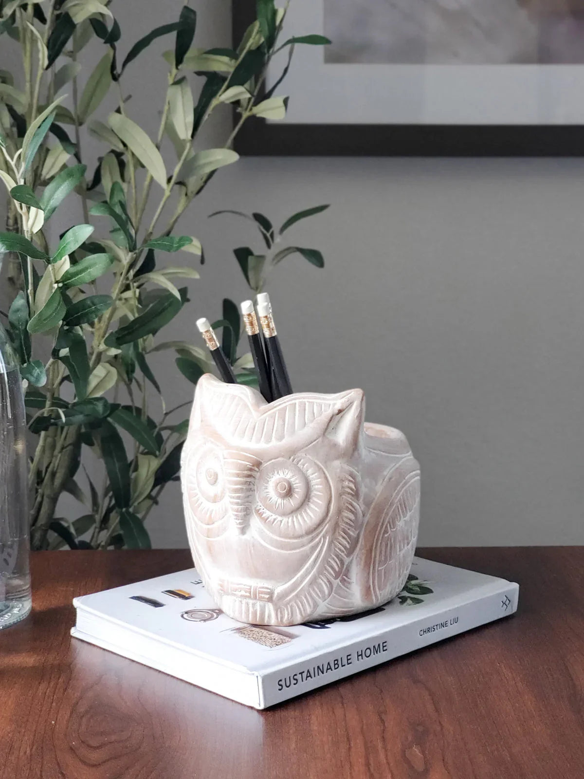 Carved terracotta owl pencil holder on a book titled Sustainable Home on a wooden table with a green plant background
