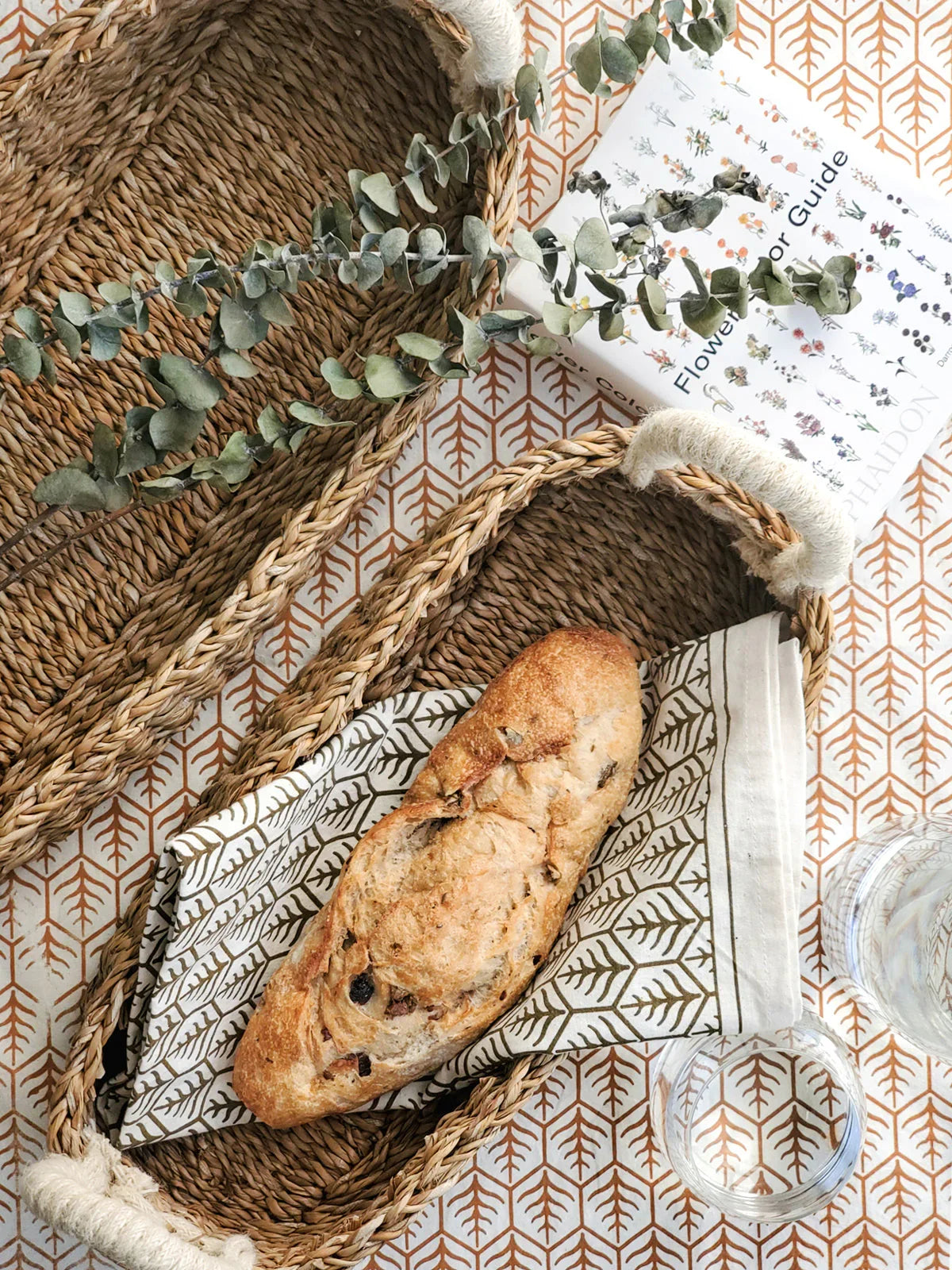 Set of two woven savar bread baskets with white handles on patterned tablecloth, one holding rustic bread and greenery