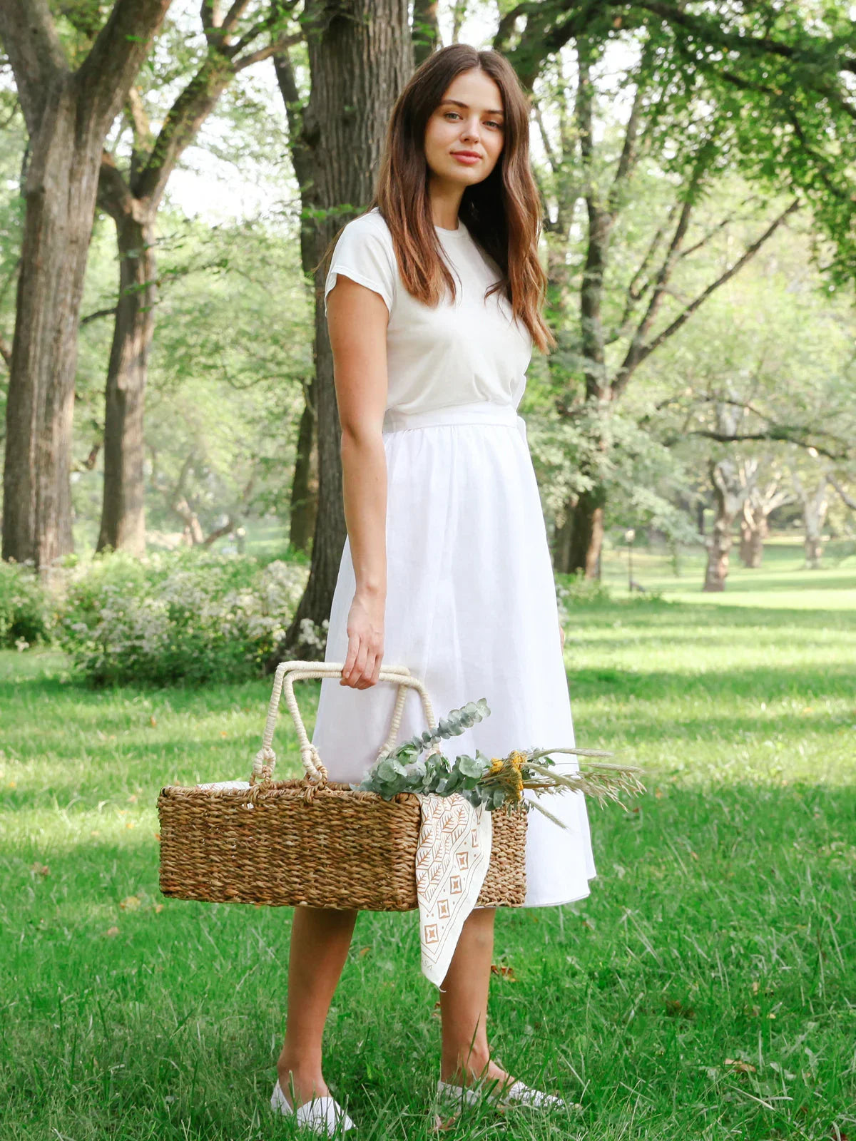 Woman in white dress holding woven picnic basket with flowers, standing in green park, Qu!rk Boutique