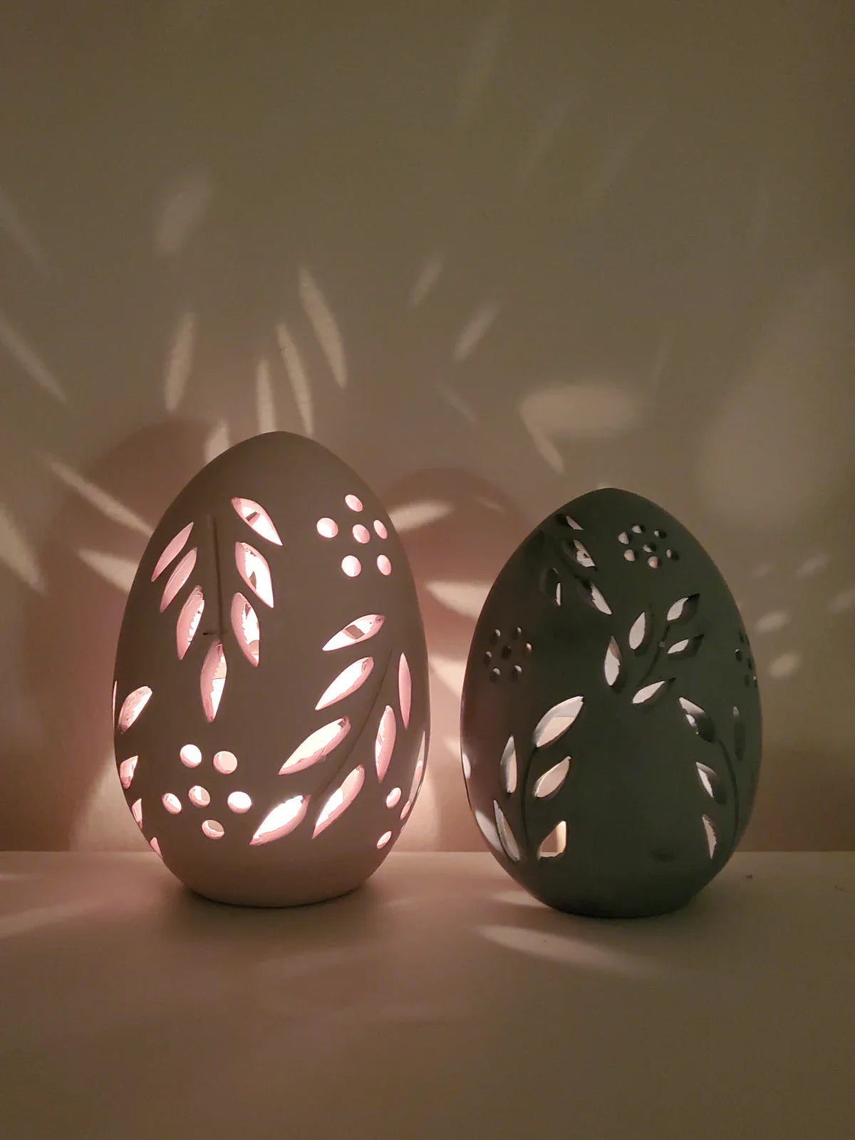 Two terracotta egg-shaped candle lanterns with leaf cutout patterns casting shadows on wall