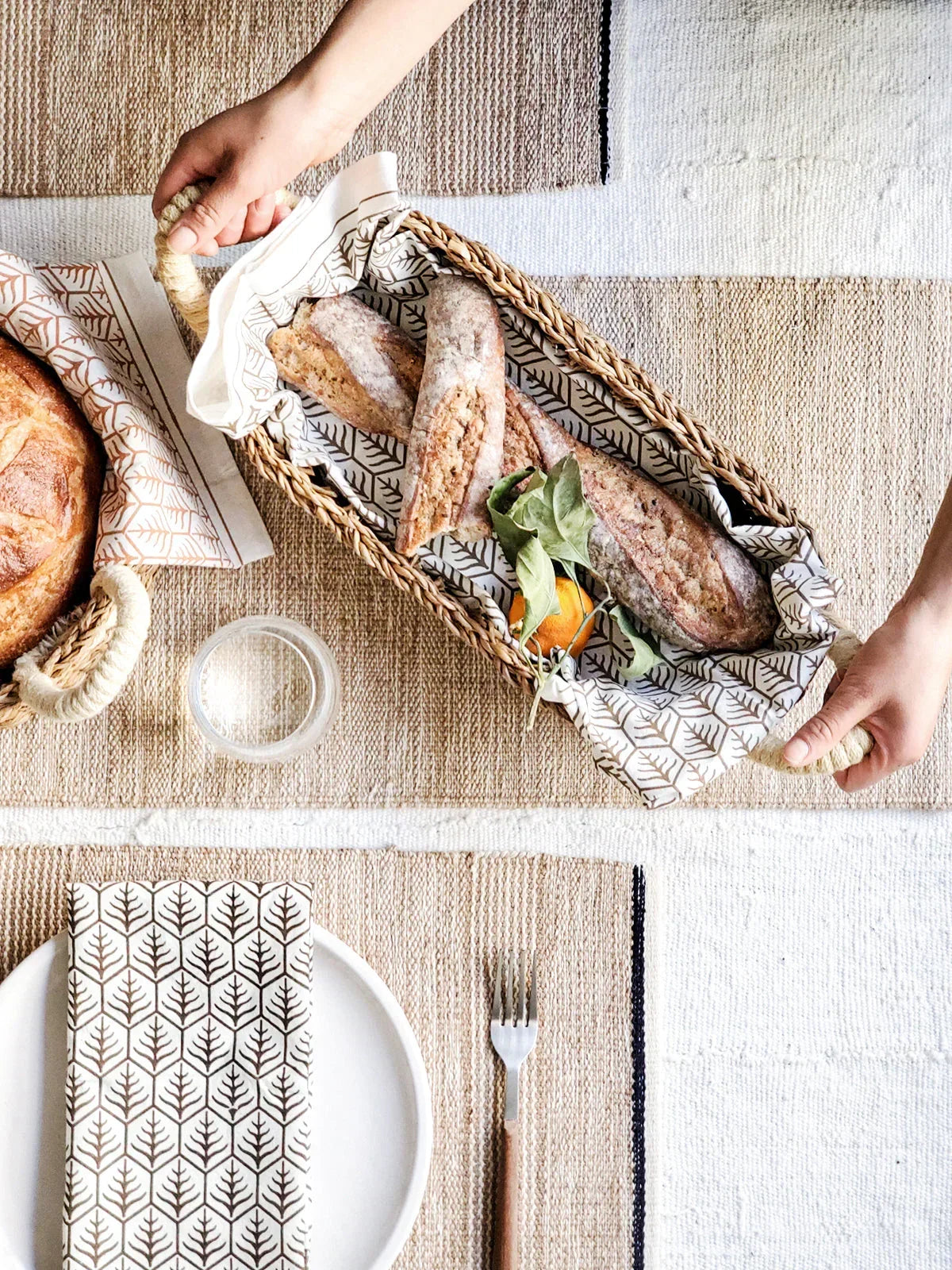 Hand holding rectangular bread basket with white handles filled with baguettes on woven table mat