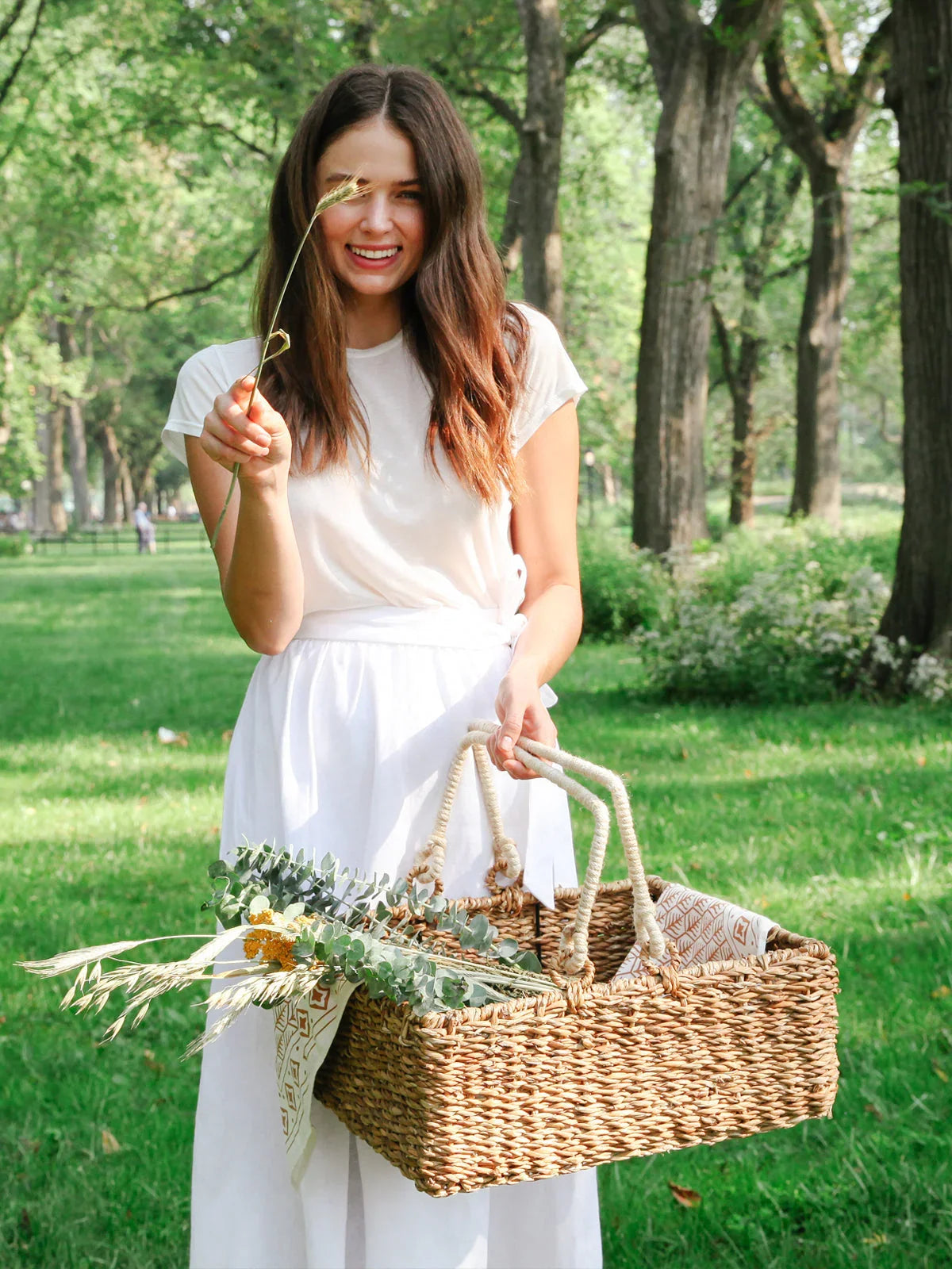 Woman in white dress holding woven picnic basket with flowers in green park, Qu!rk Boutique