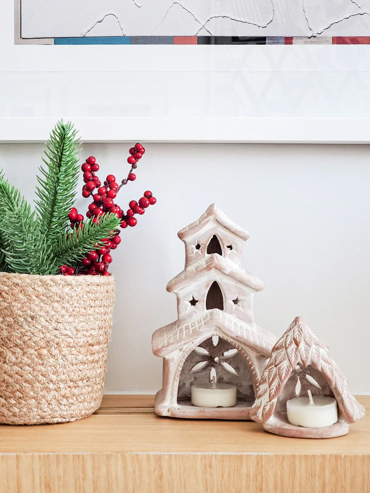 Terracotta candle holders shaped like houses with tea lights inside beside basket with pine and red berries