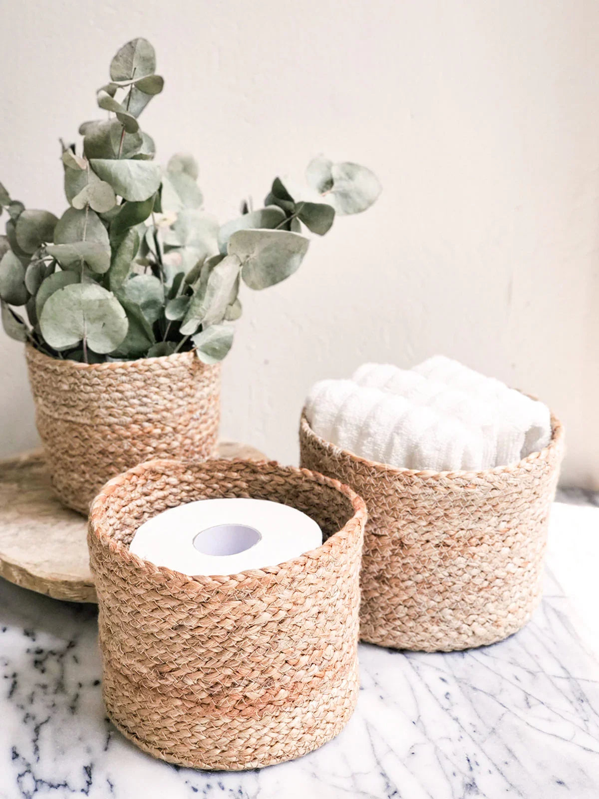 Natural woven baskets holding eucalyptus, rolled towels, and toilet paper on marble surface