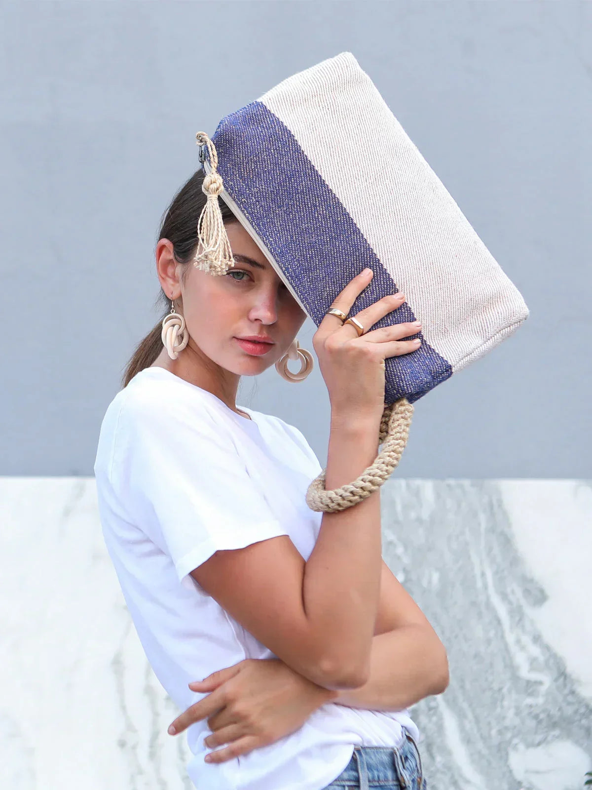Young woman wearing white shirt and blue jeans holding blue and beige woven clutch with rope handle