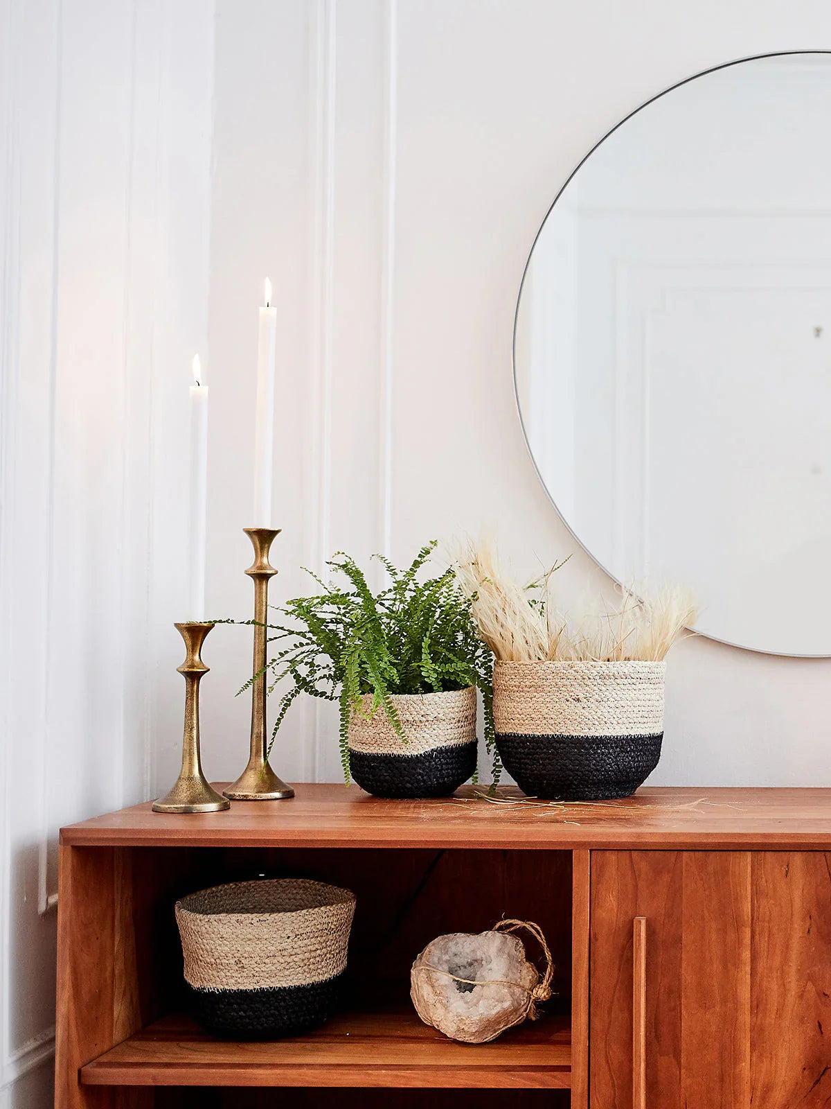 Mid-century wooden console with brass candlesticks, woven baskets, green fern, dried grasses, and round wall mirror