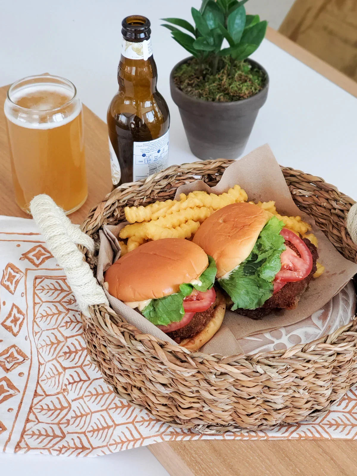 Basket with two chicken sandwiches, crinkle fries, beer bottle, beer glass, and potted plant on table