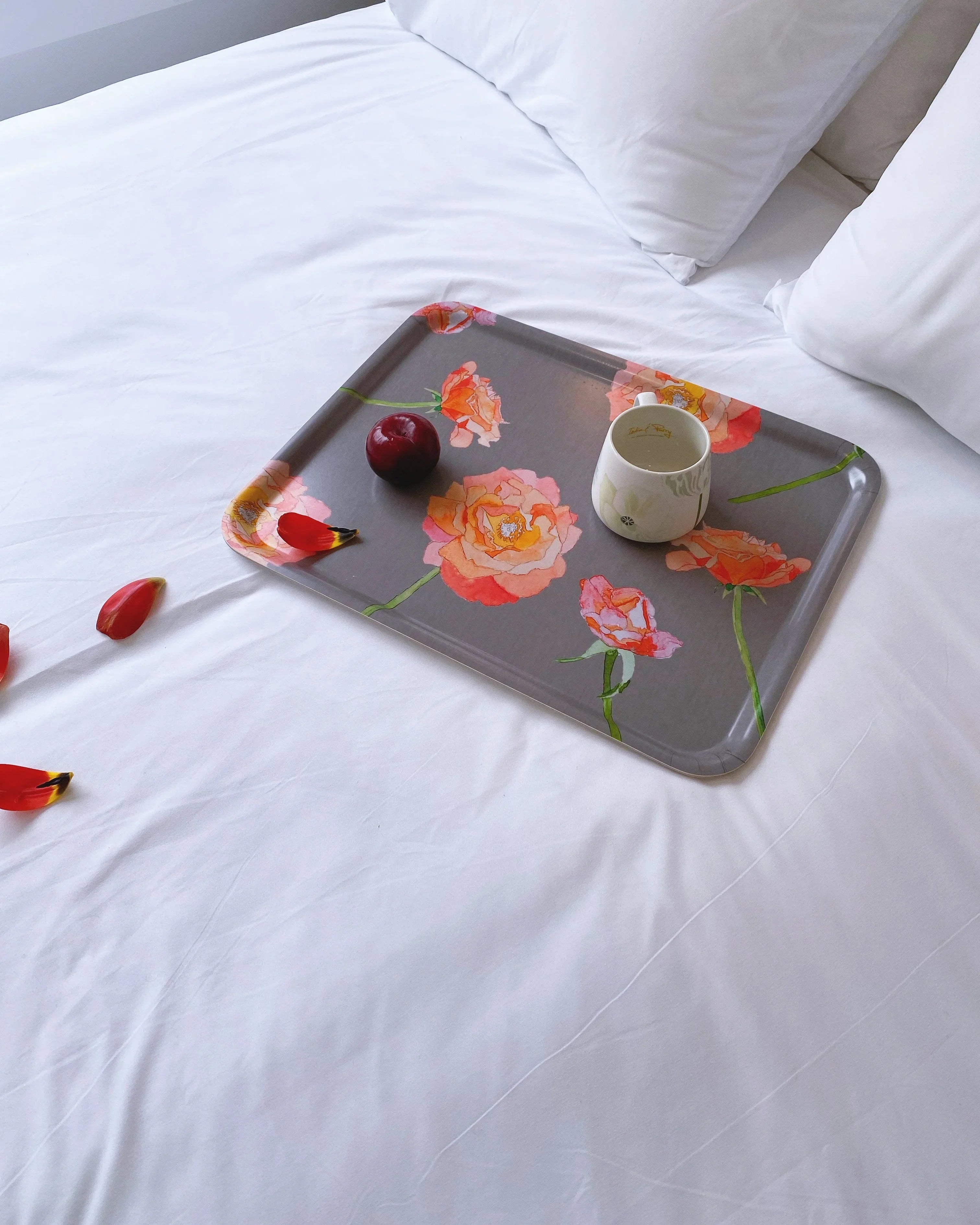 Floral tray with peach roses on white bed holding red plum and white mug, red petals nearby
