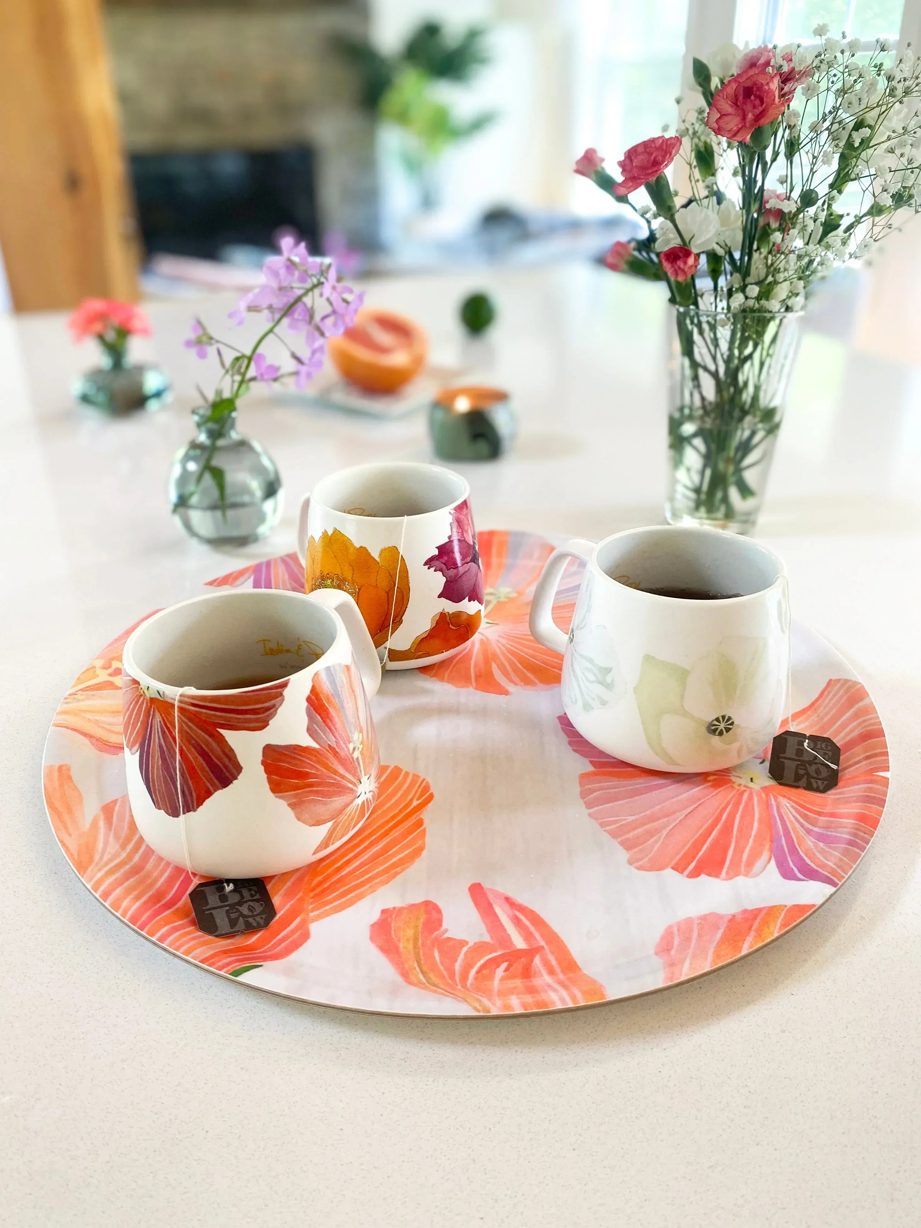 Three floral ceramic mugs with tea on a round hibiscus-printed tray on white table with flower vases in background