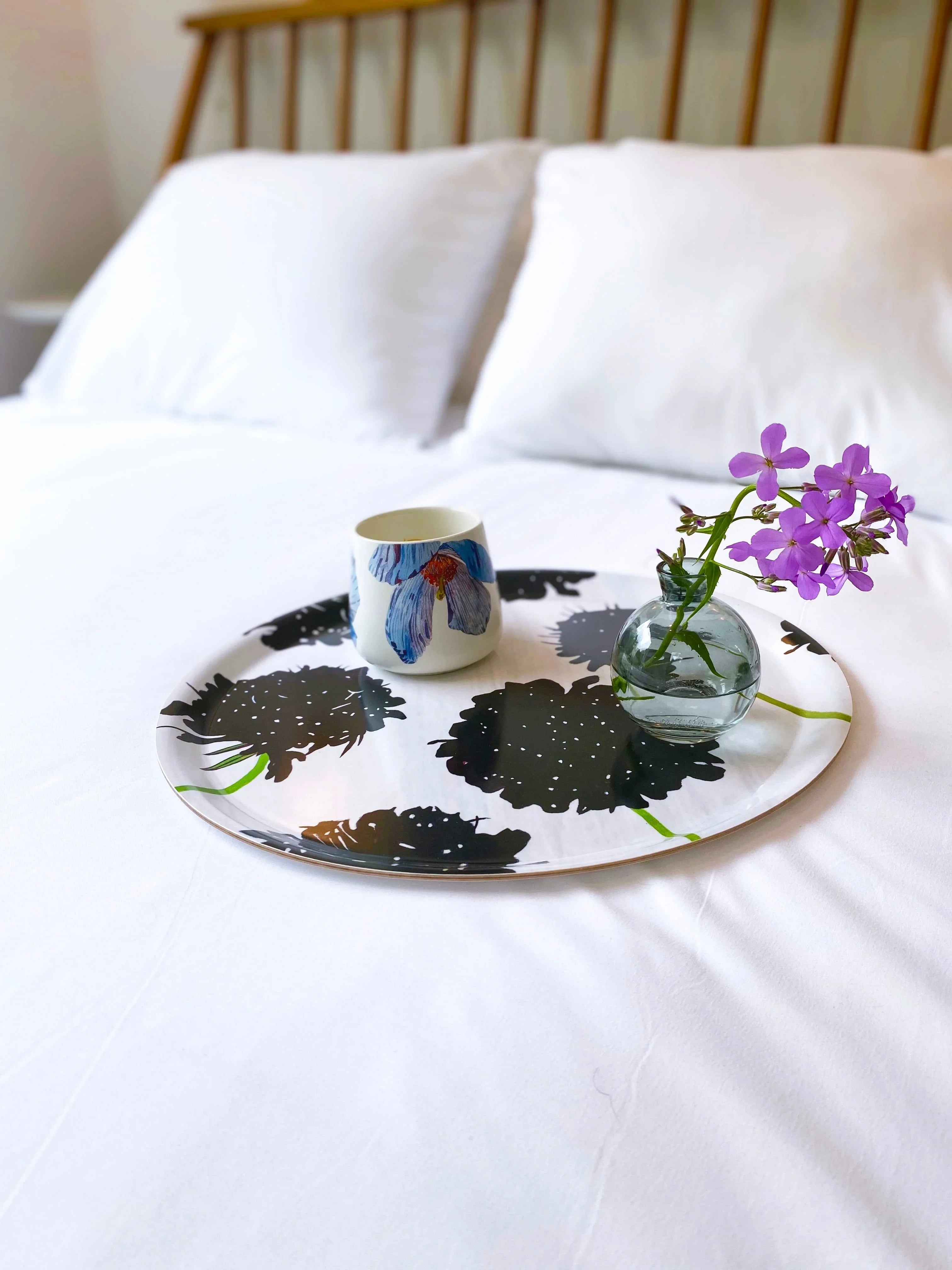 Decorative tray with black flower prints holding a blue flower mug and small vase of purple flowers on white bed