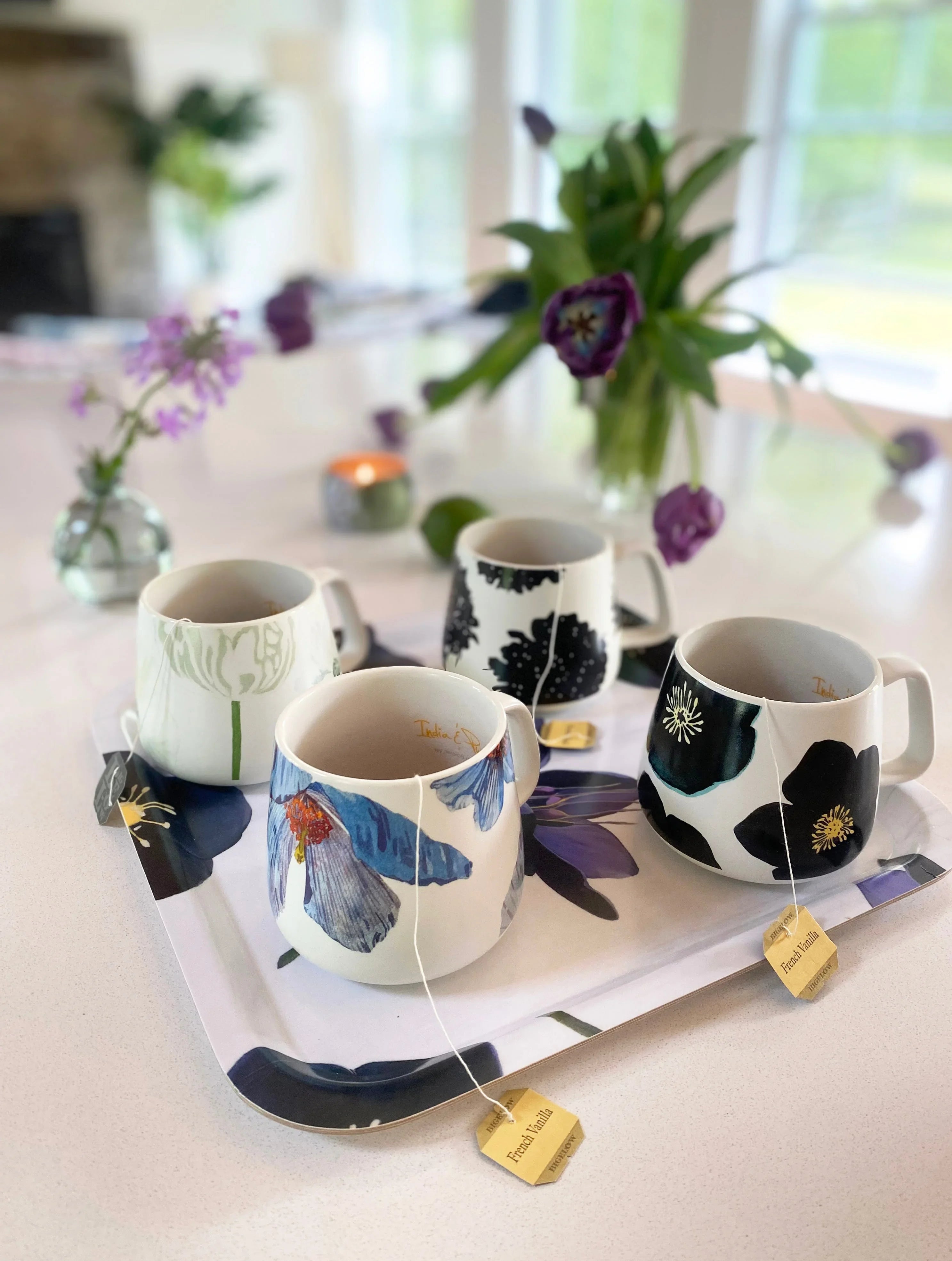 Four floral ceramic mugs with tea bags on a floral tray in a bright kitchen with purple flowers