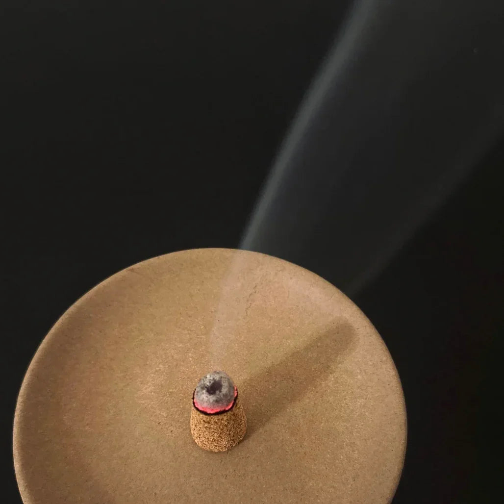 Lit incense cone on a round brown holder with smoke rising against black background