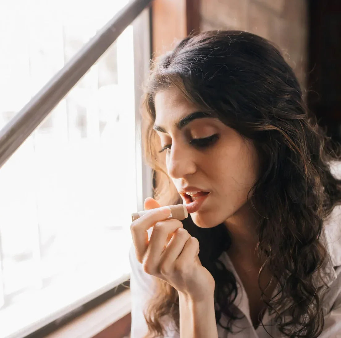Young woman applying lip balm near sunlit window with hand holding natural lipstick