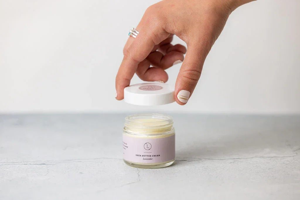 Hand lifting lid off a jar of lavender-scented shea butter cream on a light gray surface