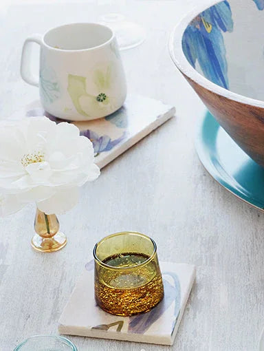 Close-up of a white poppy flower, decorative floral mug, amber glass on coasters, and bowl with blue floral interior on light table