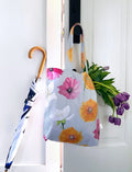 Floral tote bag with pink, orange, and white flowers hanging on a white door with purple tulips inside and a folded umbrella with wooden handle beside it
