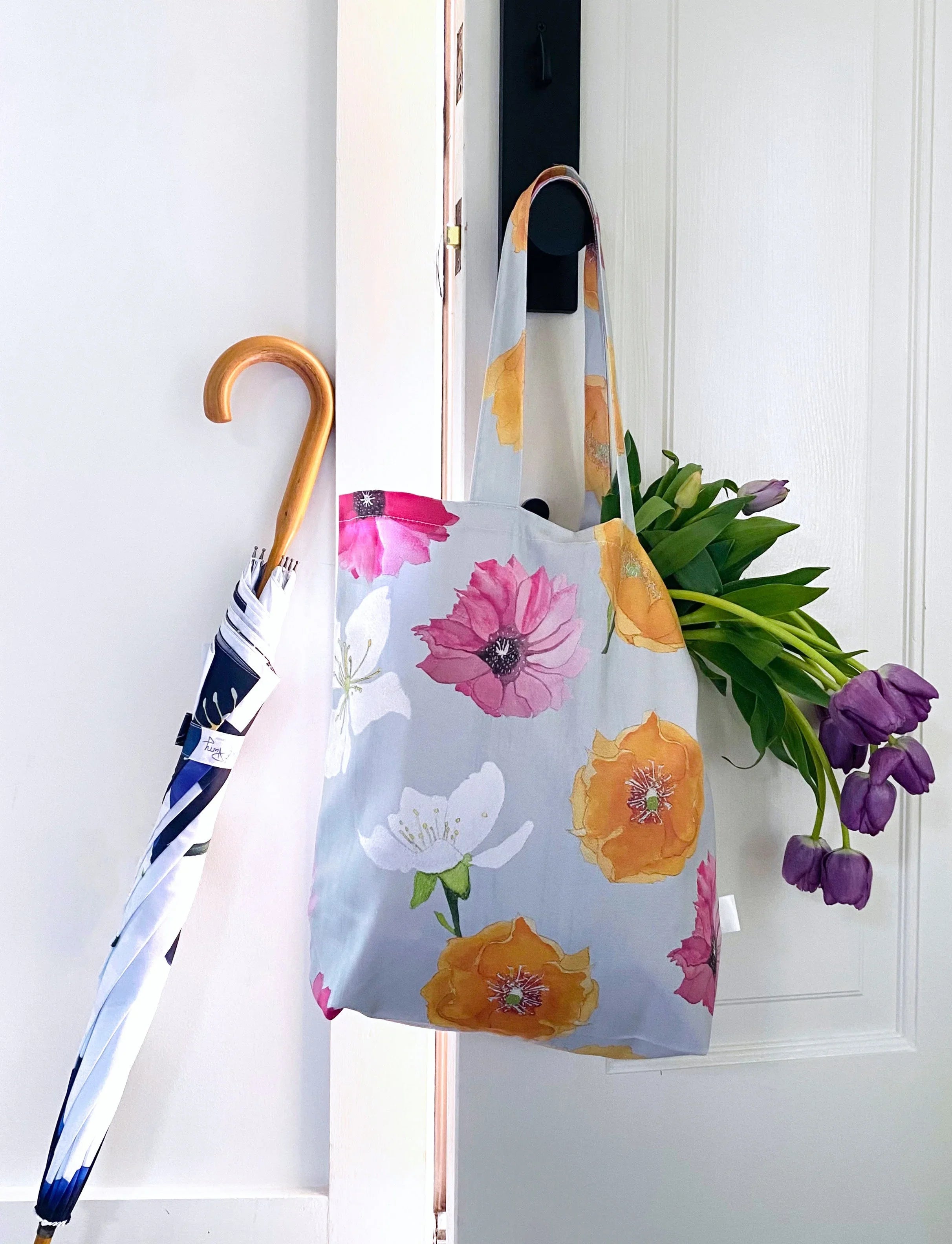 Floral tote bag with pink, orange, and white flowers hanging on a white door with purple tulips inside and a folded umbrella with wooden handle beside it