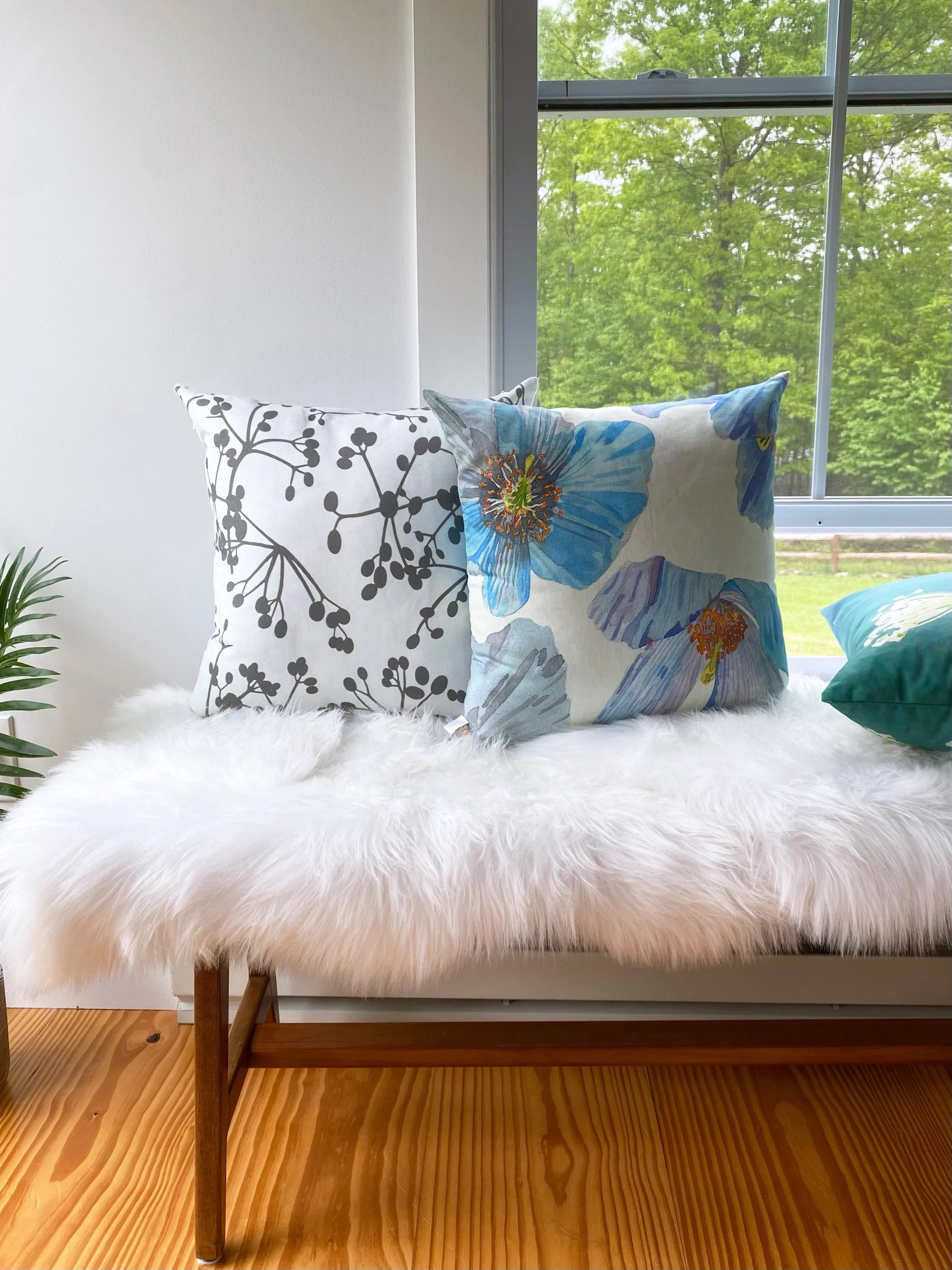 Two decorative throw pillows with viburnum and Himalayan blue poppy designs on white faux fur bench by window