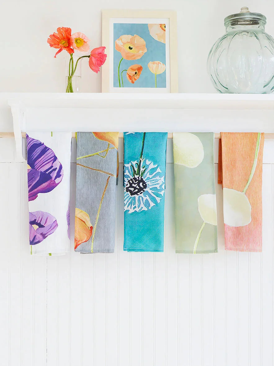 Colorful floral tea towels hanging on a white wooden rack under a shelf with flowers and framed art