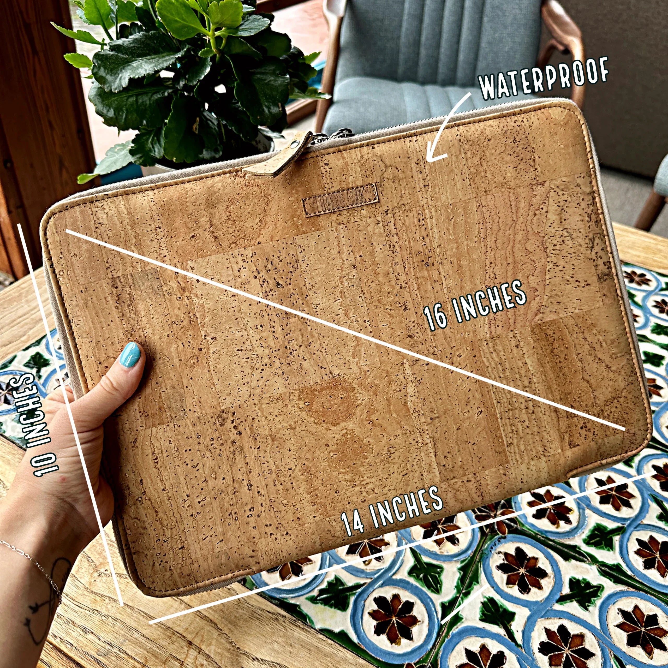 Hand holding natural cork waterproof laptop sleeve, 16 x 14 x 10 inches, on floral table.