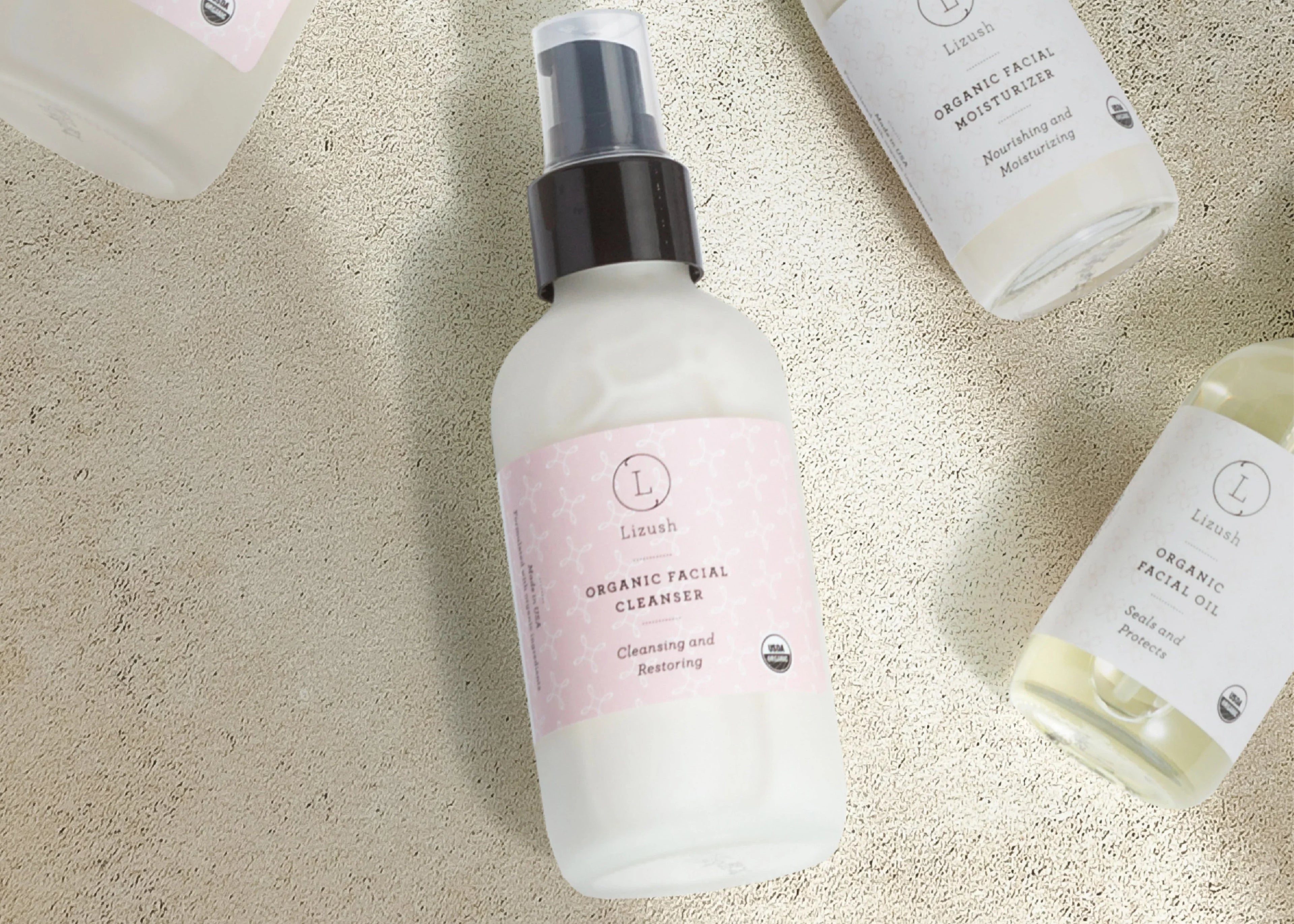 Bottle of Lizush Organic Facial Cleanser with black pump on textured surface, surrounded by moisturizer and facial oil bottles