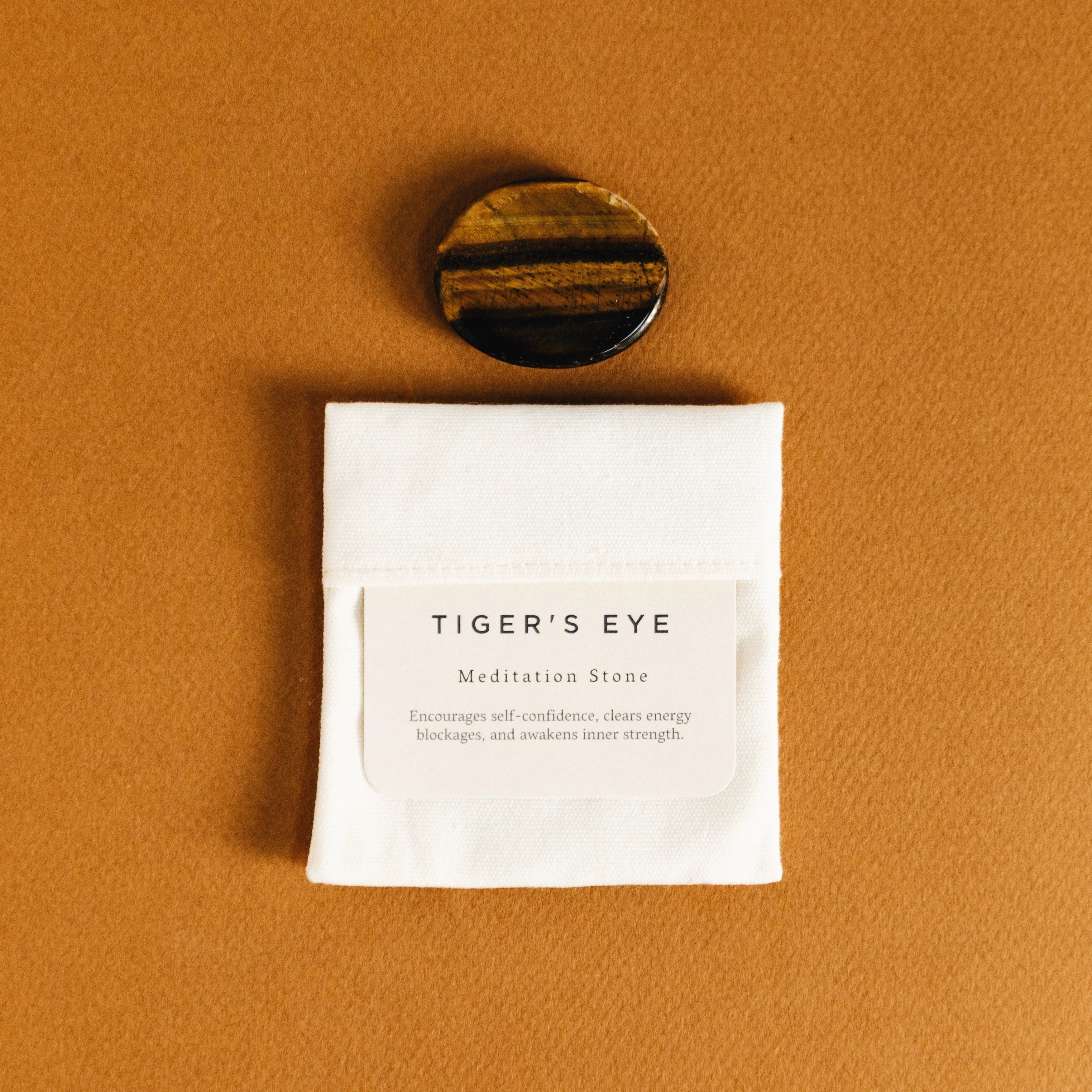 Polished tiger's eye meditation stone on a brown surface with a white pouch and label