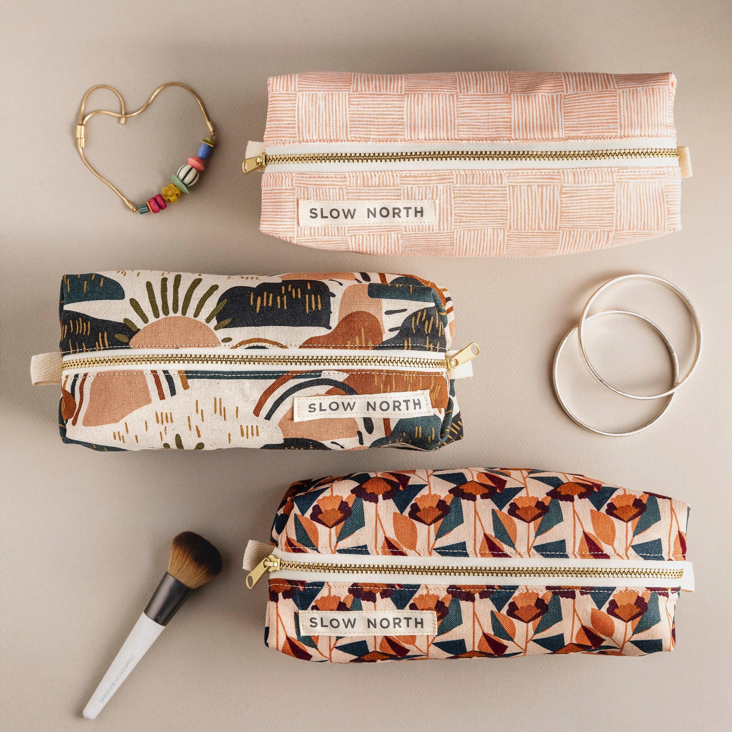 Three patterned Slow North travel pouches with gold zippers on beige background, accompanied by jewelry and makeup brush