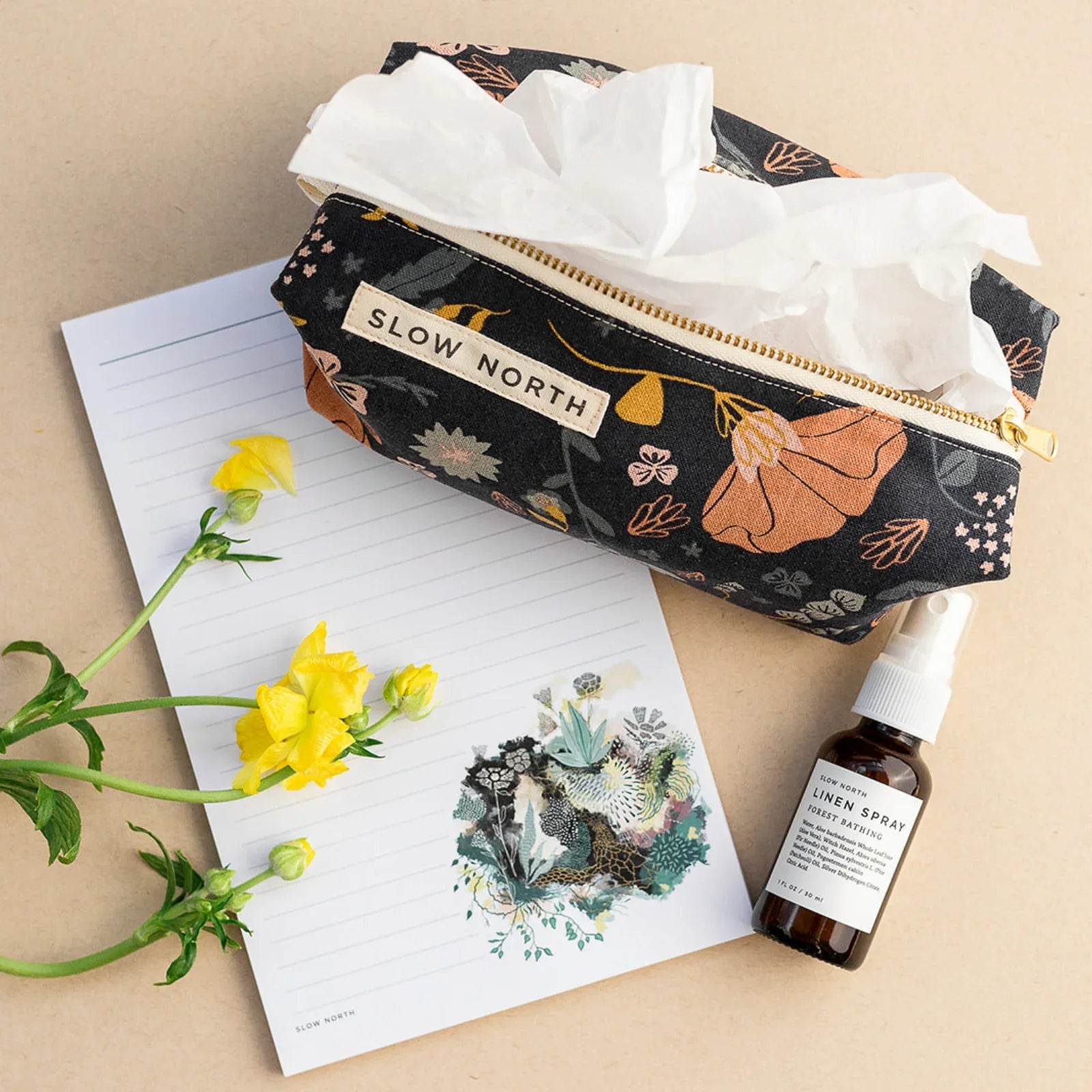 Slow North floral travel pouch with tissues, lined paper with botanical design, and linen spray bottle on beige background