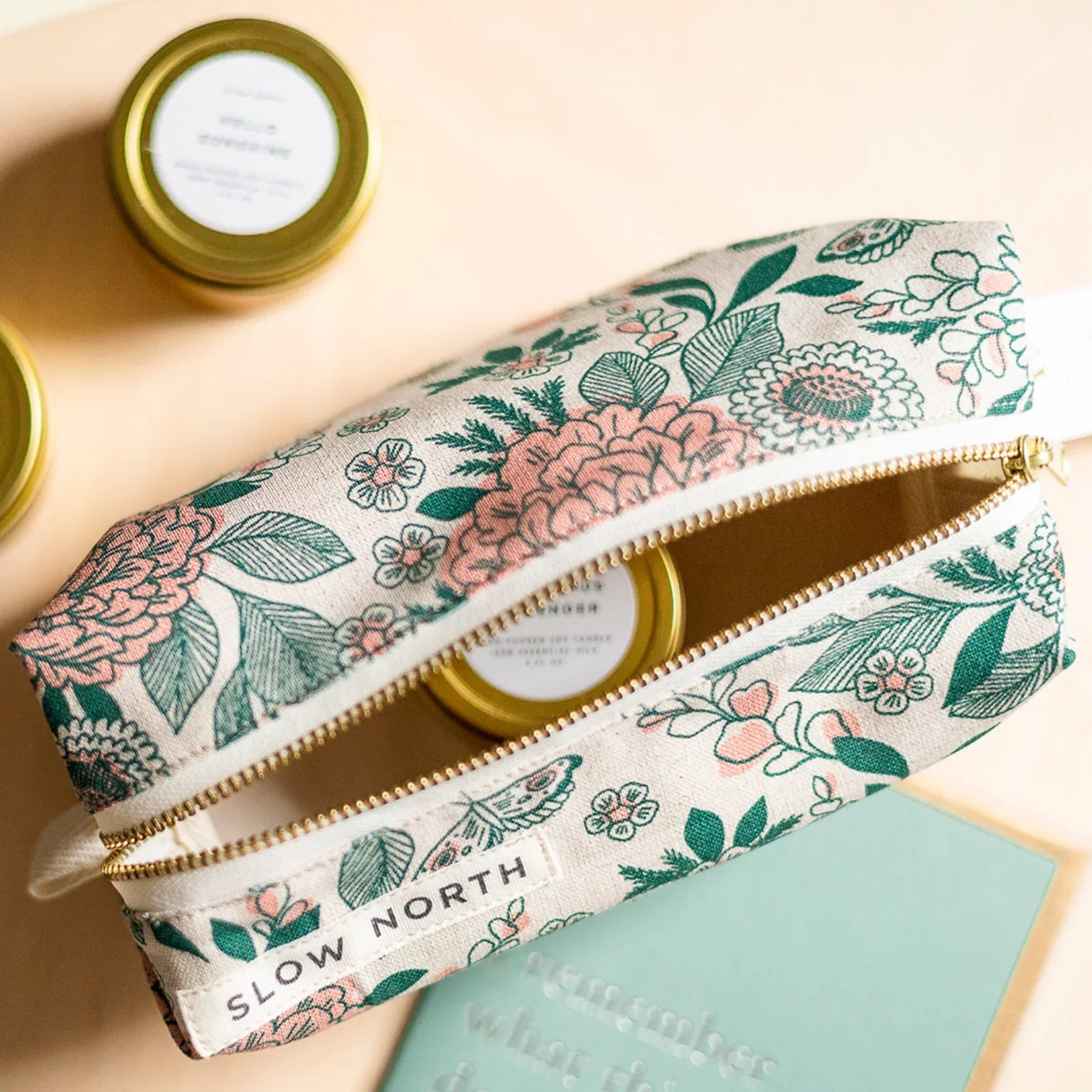 Floral travel pouch with gold zipper and Slow North label, holding small skincare tins on a peach background