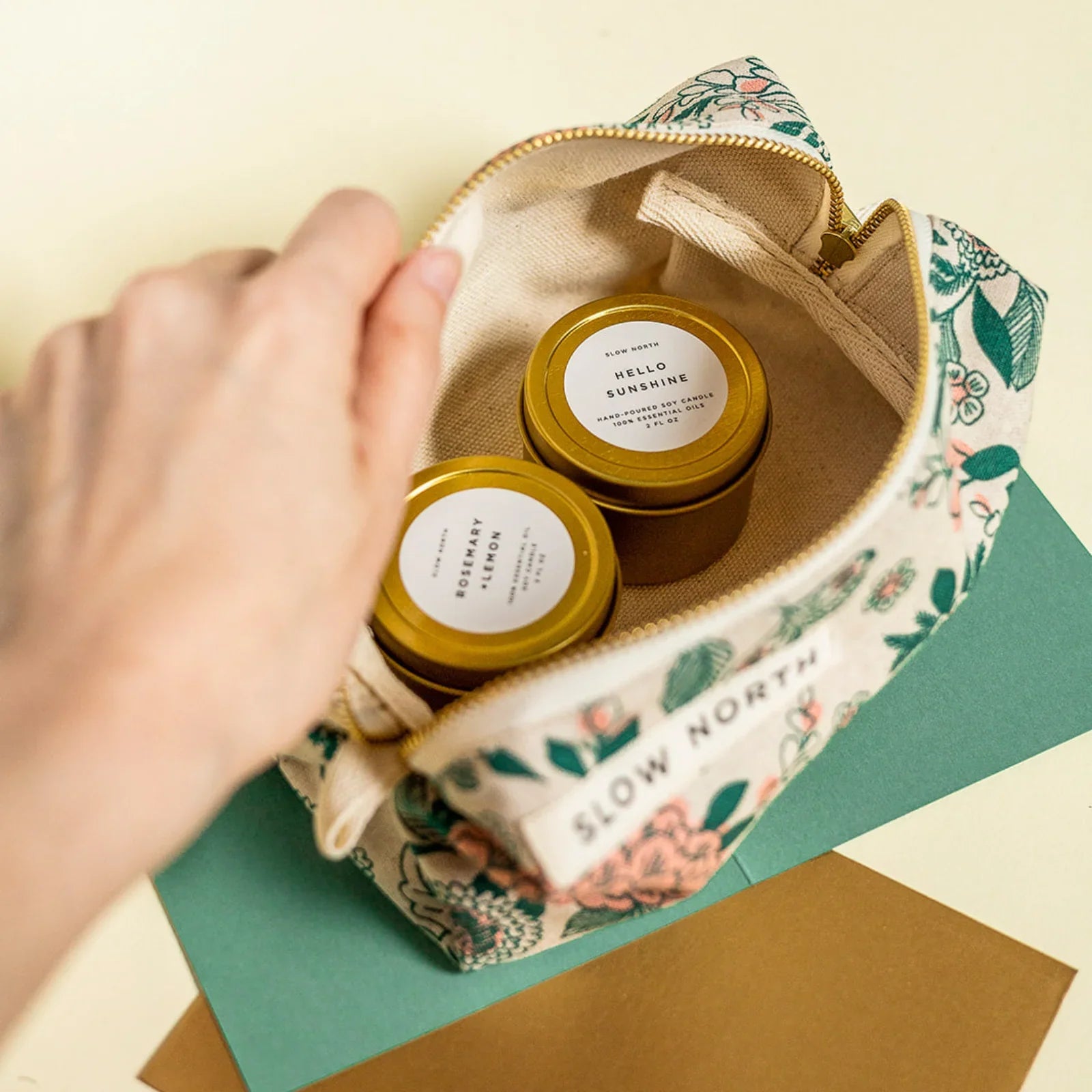 Hand opening floral travel pouch with two gold-tin soy candles labeled Hello Sunshine and Rosemary Lemon