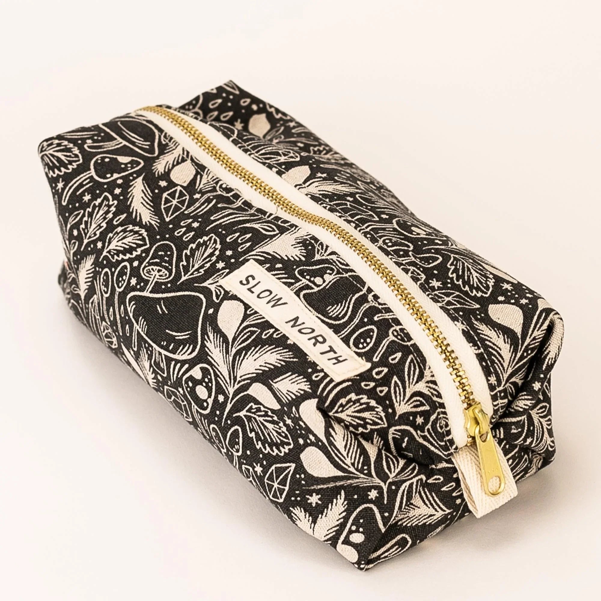Black fabric travel pouch with white mystical mushroom and leaf pattern, gold zipper, Slow North label