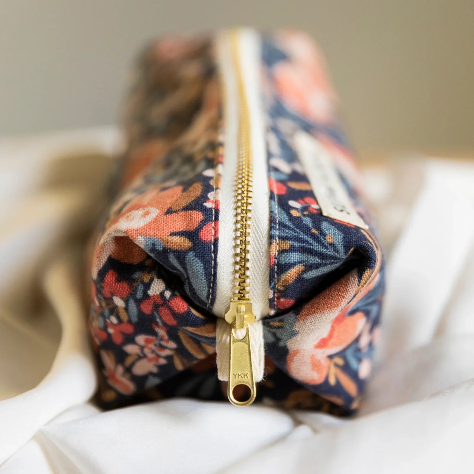 Close-up of a floral travel pouch with navy, pink, and orange pattern and gold YKK zipper on white fabric