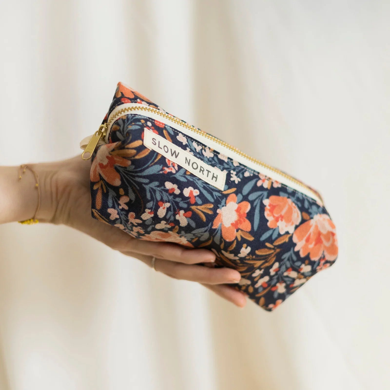Hand holding a floral travel pouch with orange, pink flowers and Slow North label