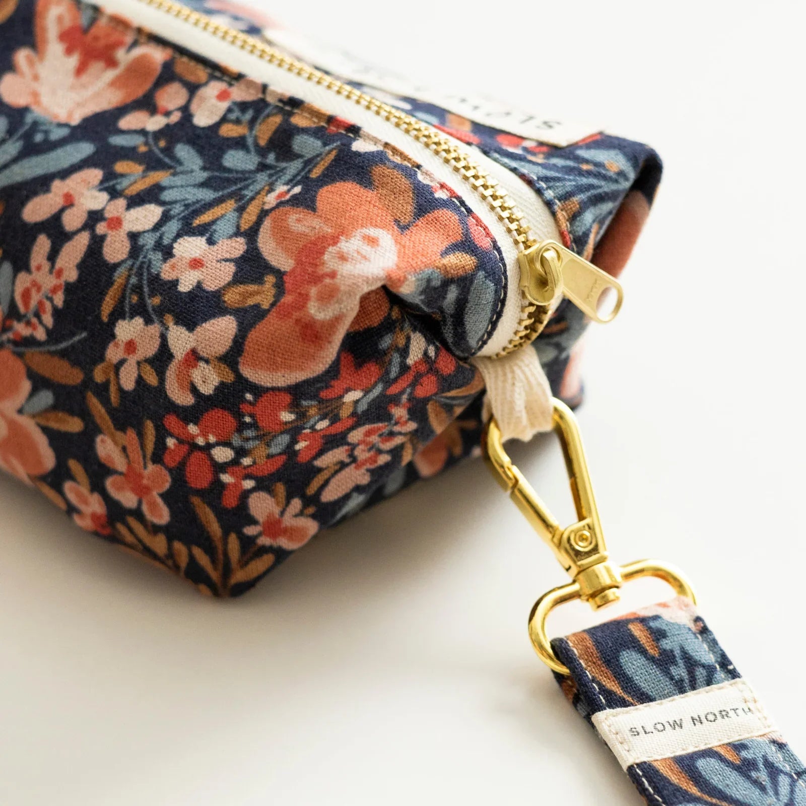 Close-up of a floral travel pouch with gold zipper and detachable strap labeled Slow North
