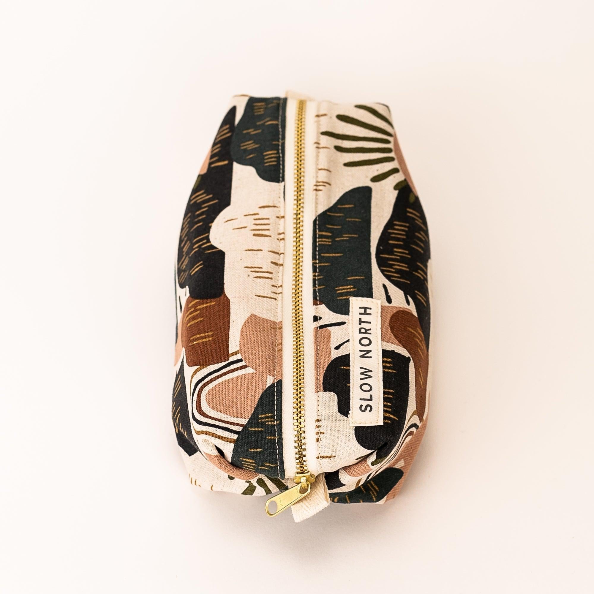 Rectangular travel pouch with abstract earth tone pattern and gold zipper, labeled Slow North