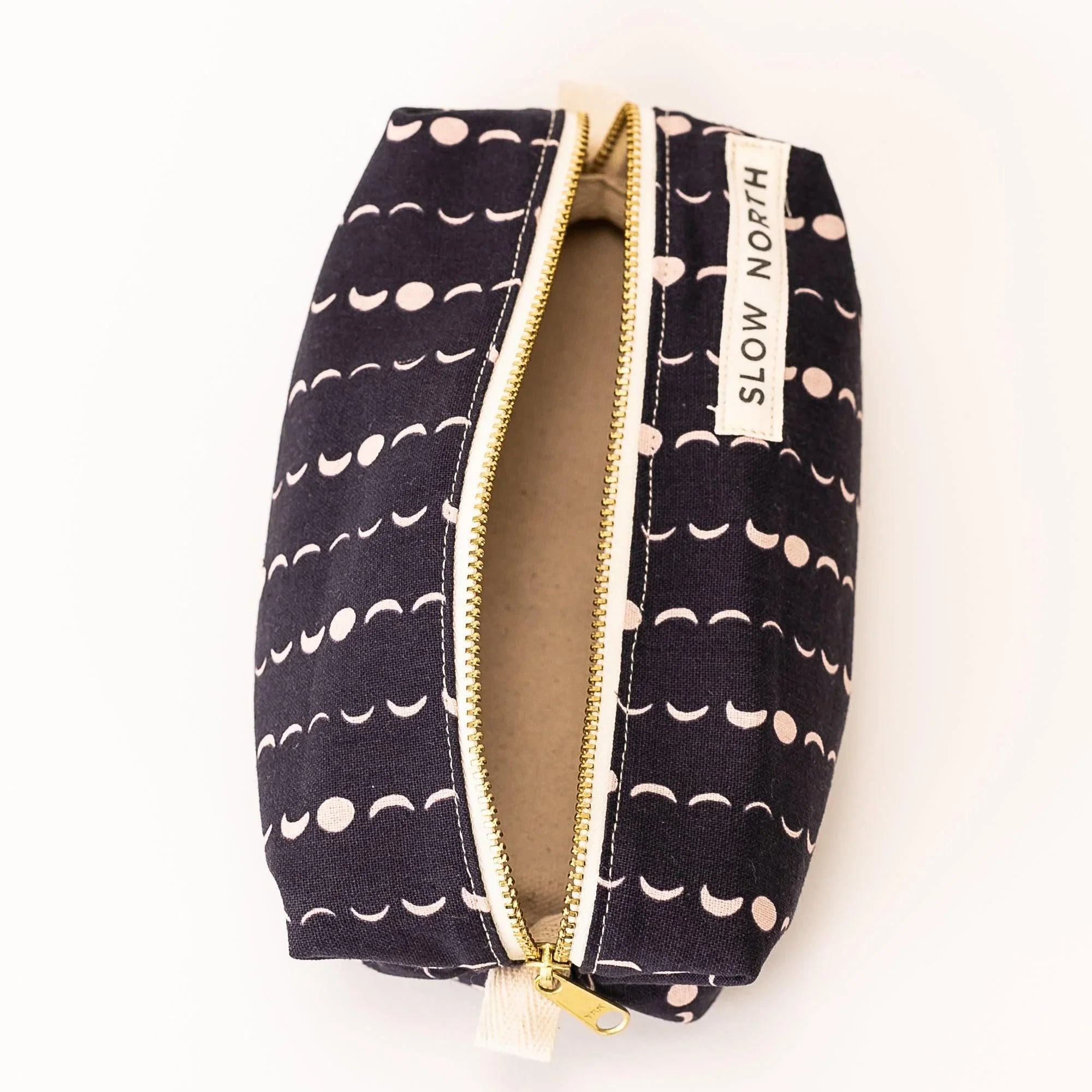 Black travel pouch with white moon phase pattern and gold zipper, open showing beige interior, Slow North label