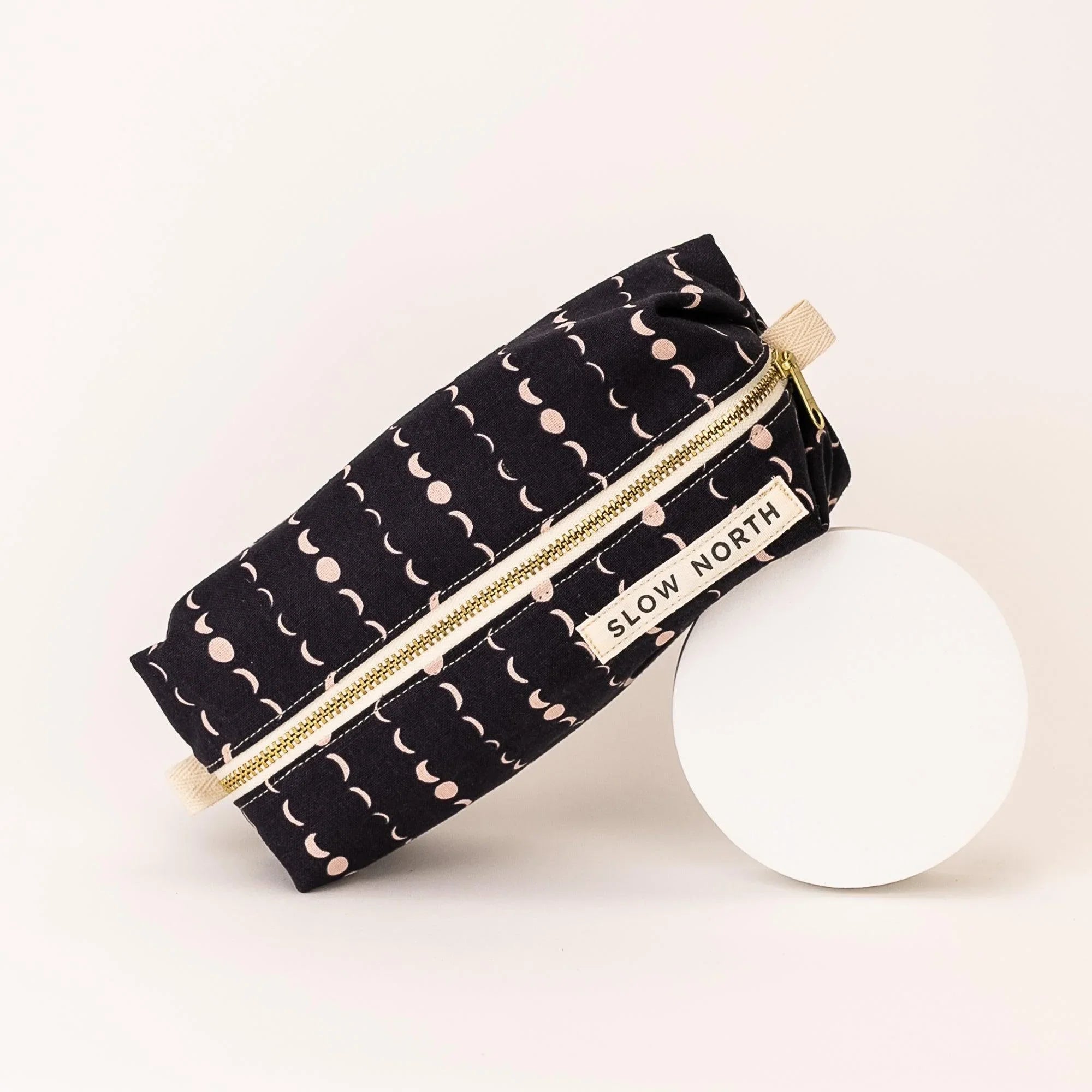 Black travel pouch with cream moon phase pattern and gold zipper, branded Slow North, on white background