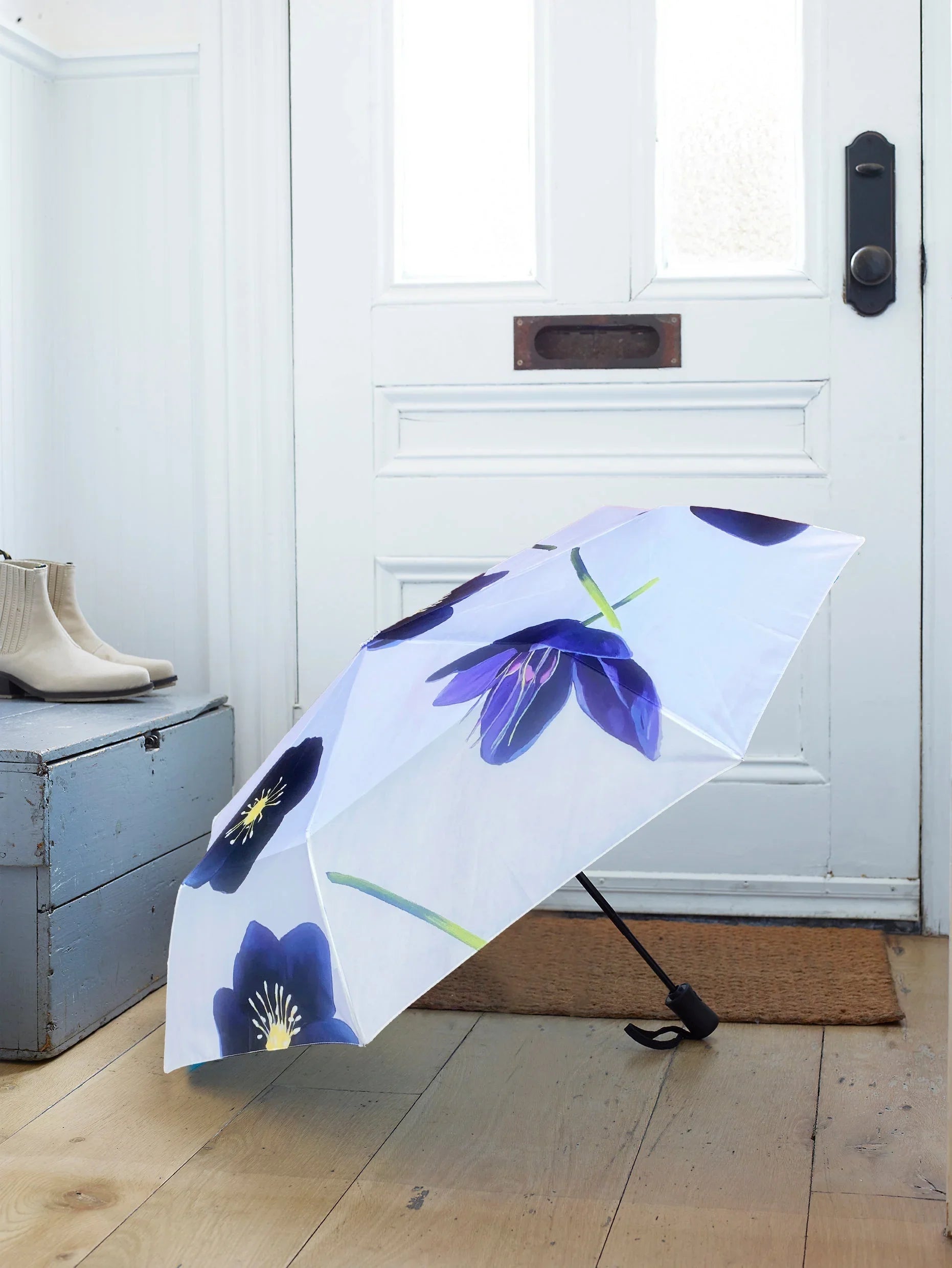 Blue hellebore flower patterned umbrella open on wooden floor near white front door and gray bench with beige boots