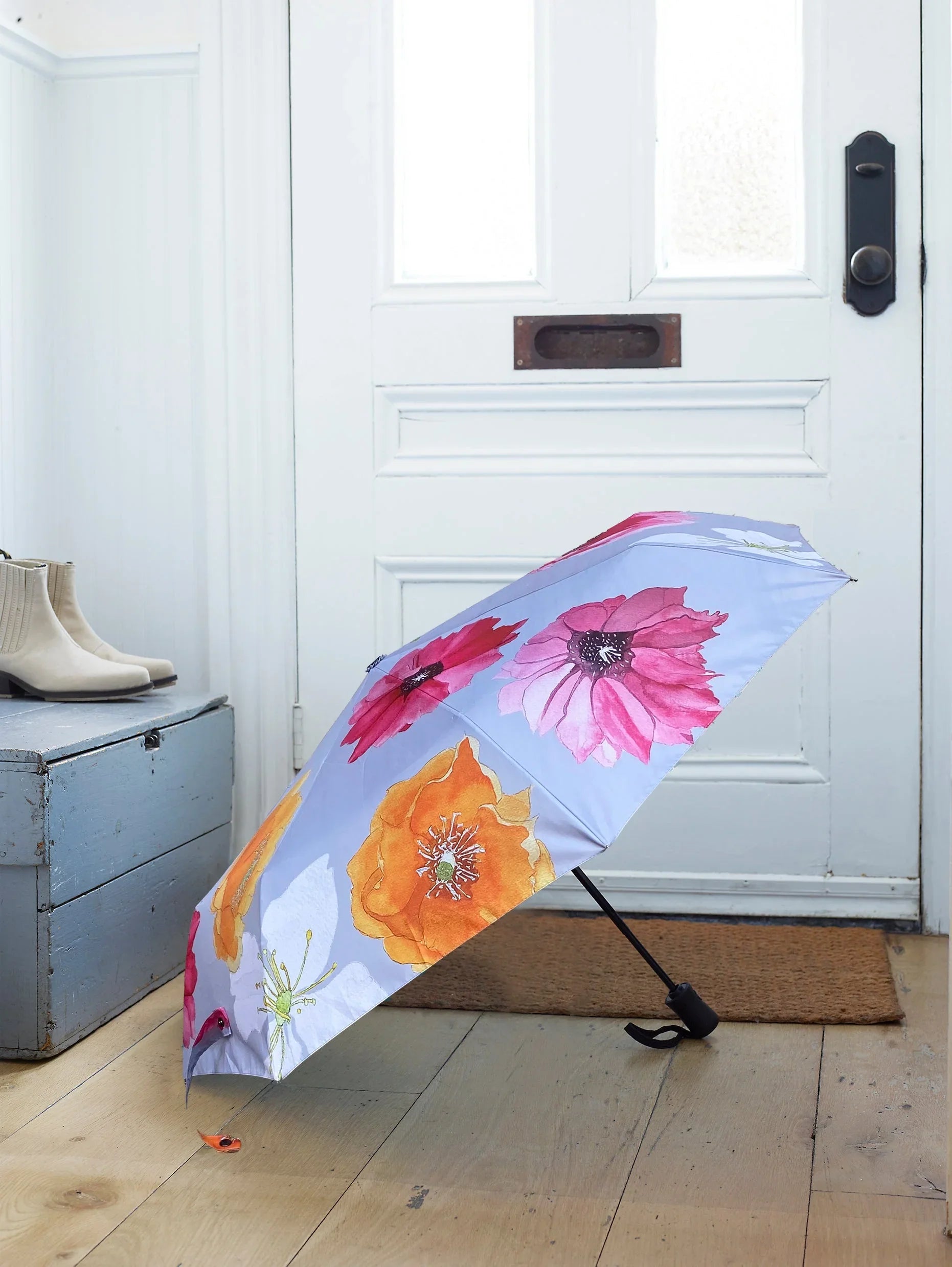 Folded umbrella with bright pink and orange flower print on wooden floor by white door and blue wooden chest