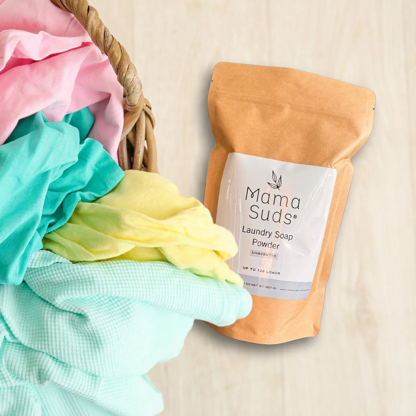 Mama Suds laundry soap powder and colorful folded clothes in a basket on a light wood surface