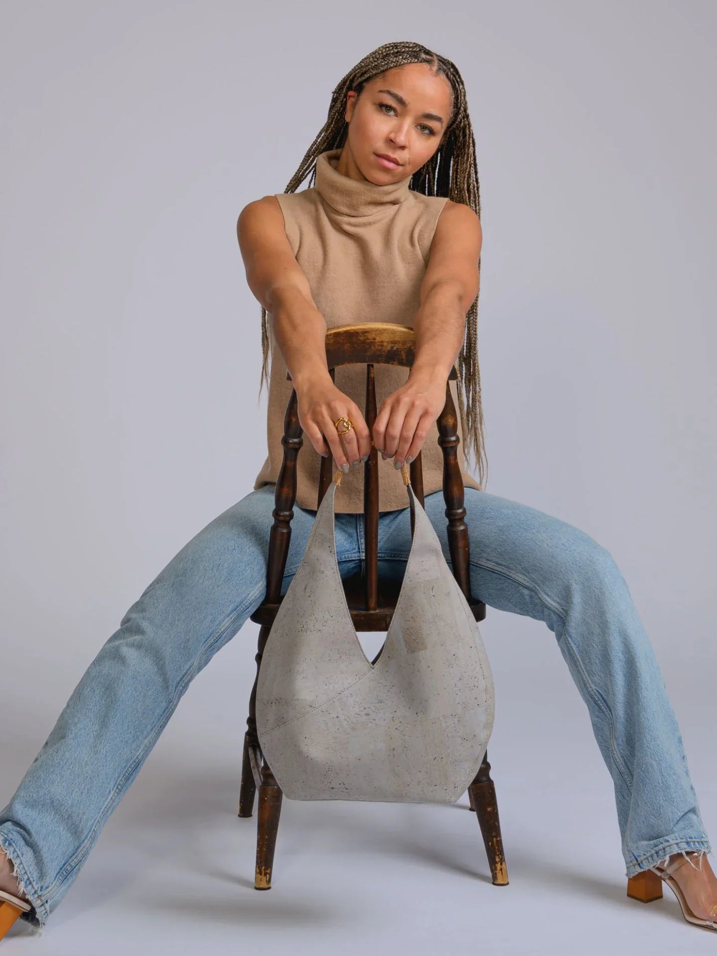 Woman with long braids in a beige sleeveless turtleneck and jeans posing on a chair, holding a sustainable grey hobo bag from Qu!rk Boutique.