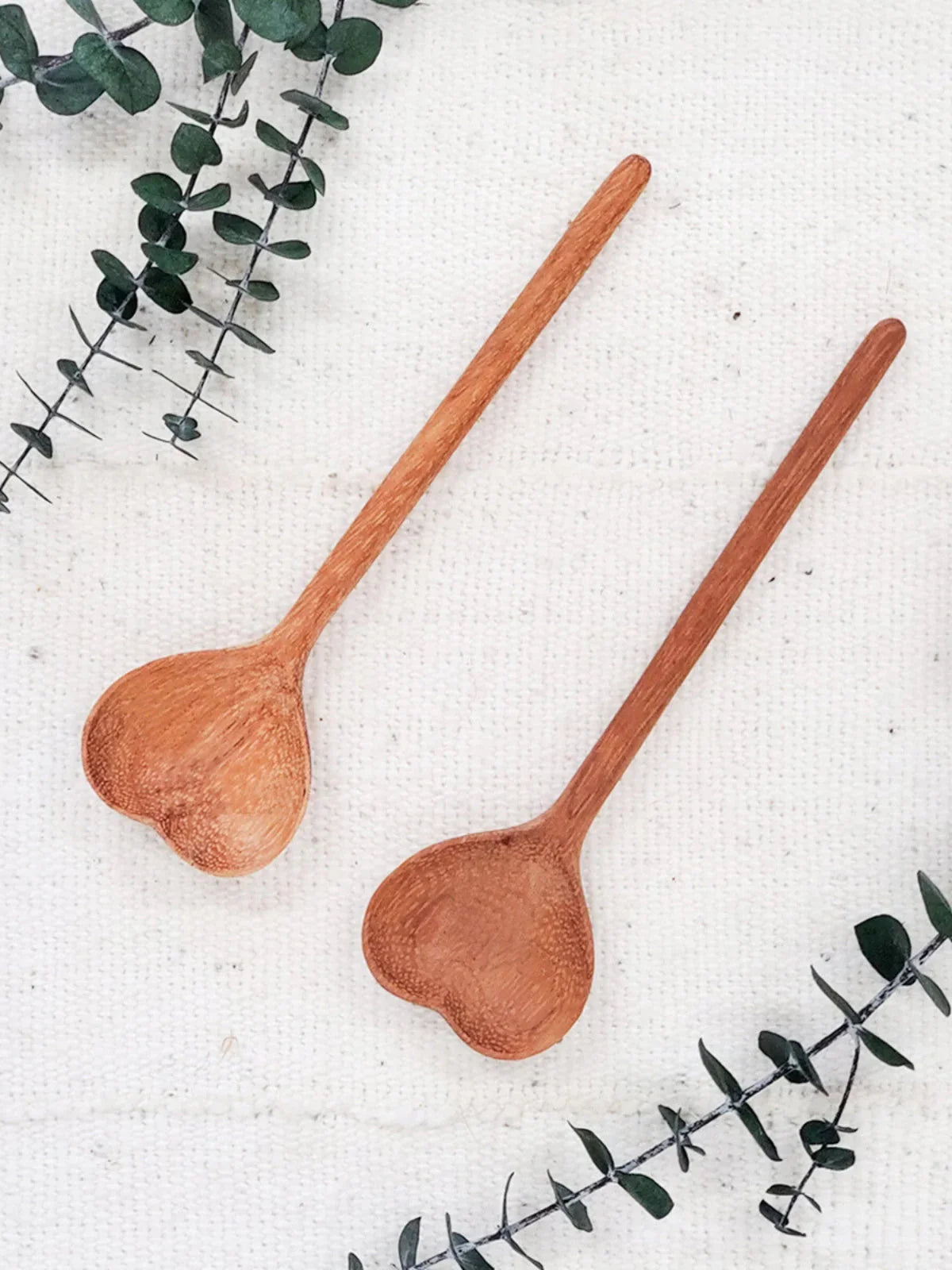 Two wooden heart-shaped spoons on white textured fabric with green eucalyptus leaves