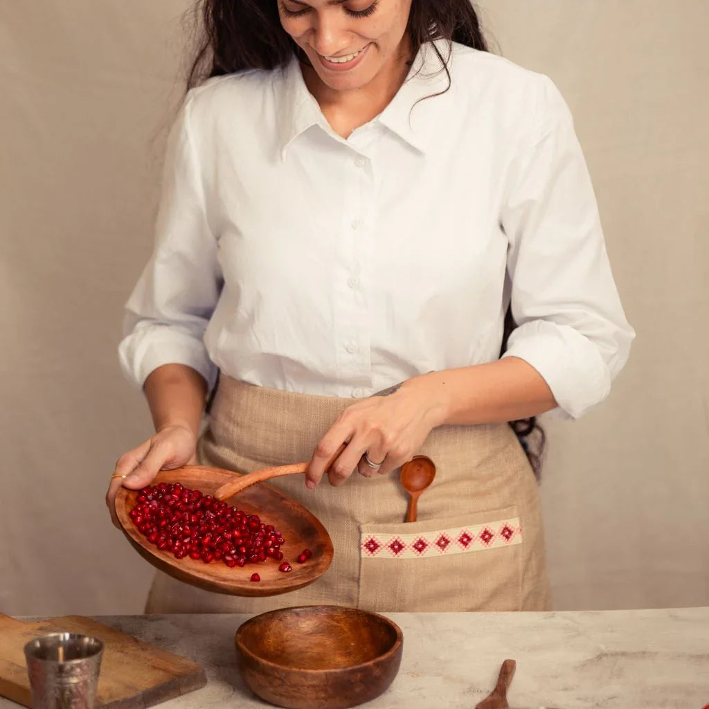 Woman in white shirt and linen apron pours pomegranate seeds into wooden bowl, artisan kitchen