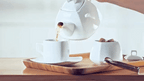 Pouring tea from modern white teapot into cup on wooden tray, styled for Qu!rk Boutique gifts