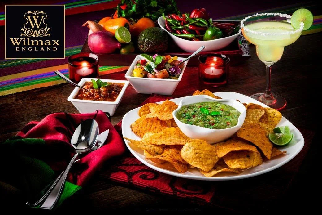 Colorful Mexican-inspired appetizer spread with tortilla chips, guacamole, salsa, fresh limes, and a margarita on a dark table, featuring Wilmax England serveware.